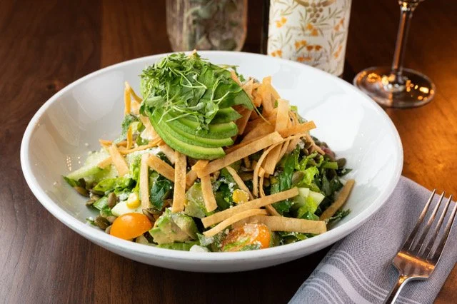 Salad with greens, avocado slices, shredded cheese, cherry tomatoes, served in a white bowl on a wooden table, with a fork and napkin nearby.