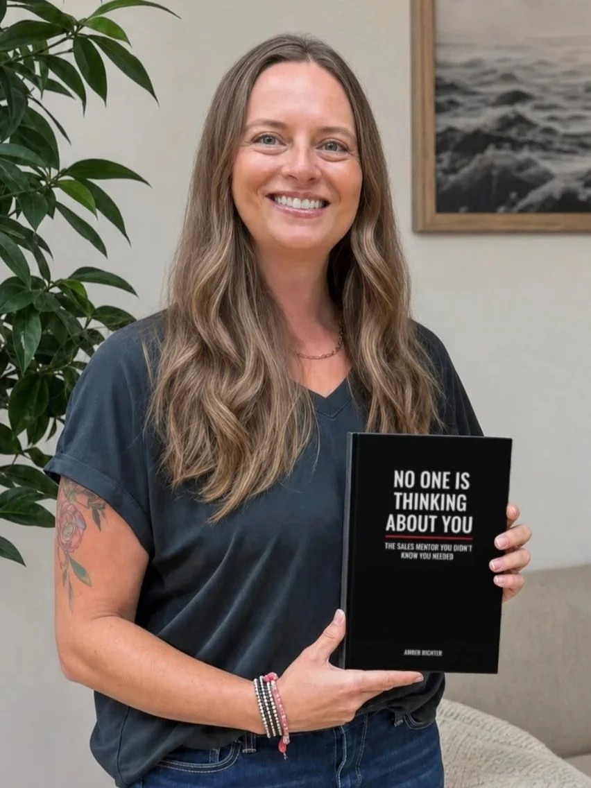 A smiling woman with long wavy hair holding a black book titled 'No One Is Thinking About You' in an office setting with a large plant and framed picture of waves on the wall.