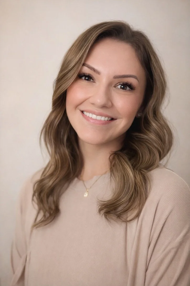 A woman with light brown, wavy hair wearing a beige top and a gold necklace, smiling at the camera against a plain background.