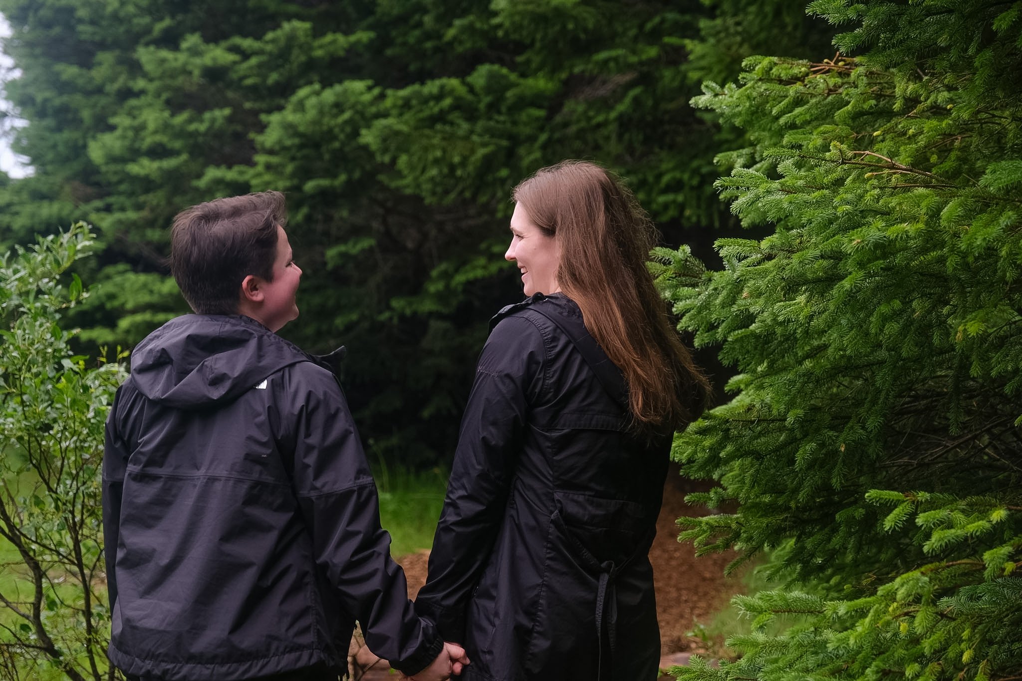 A man and a woman holding hands and smiling at each other outdoors surrounded by green trees and bushes.