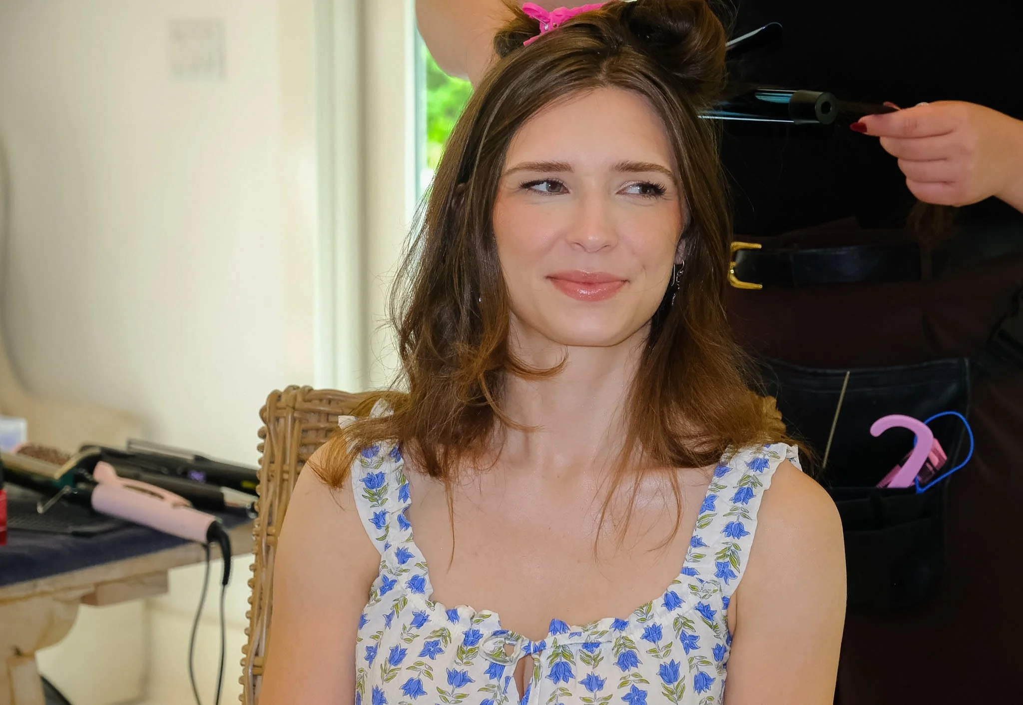 A woman with reddish-brown hair and light skin sits in a wicker chair at a vanity mirror, getting her hair styled. She wears a white dress with blue floral pattern and has a slight smile on her face.