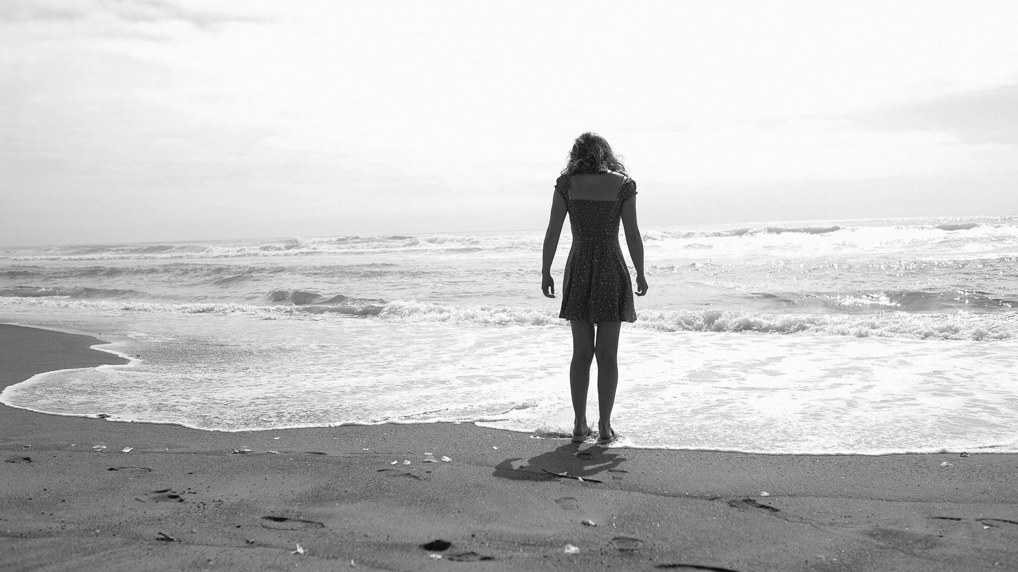 A black-and-white photo of a woman standing on the beach, looking at the ocean waves.
