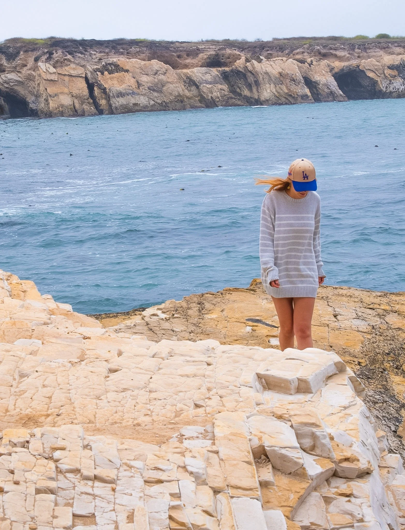 A young woman wearing a beige Los Angeles Dodgers cap and a light-colored, striped sweater stands on rocks by the ocean, with rocky cliffs in the background.