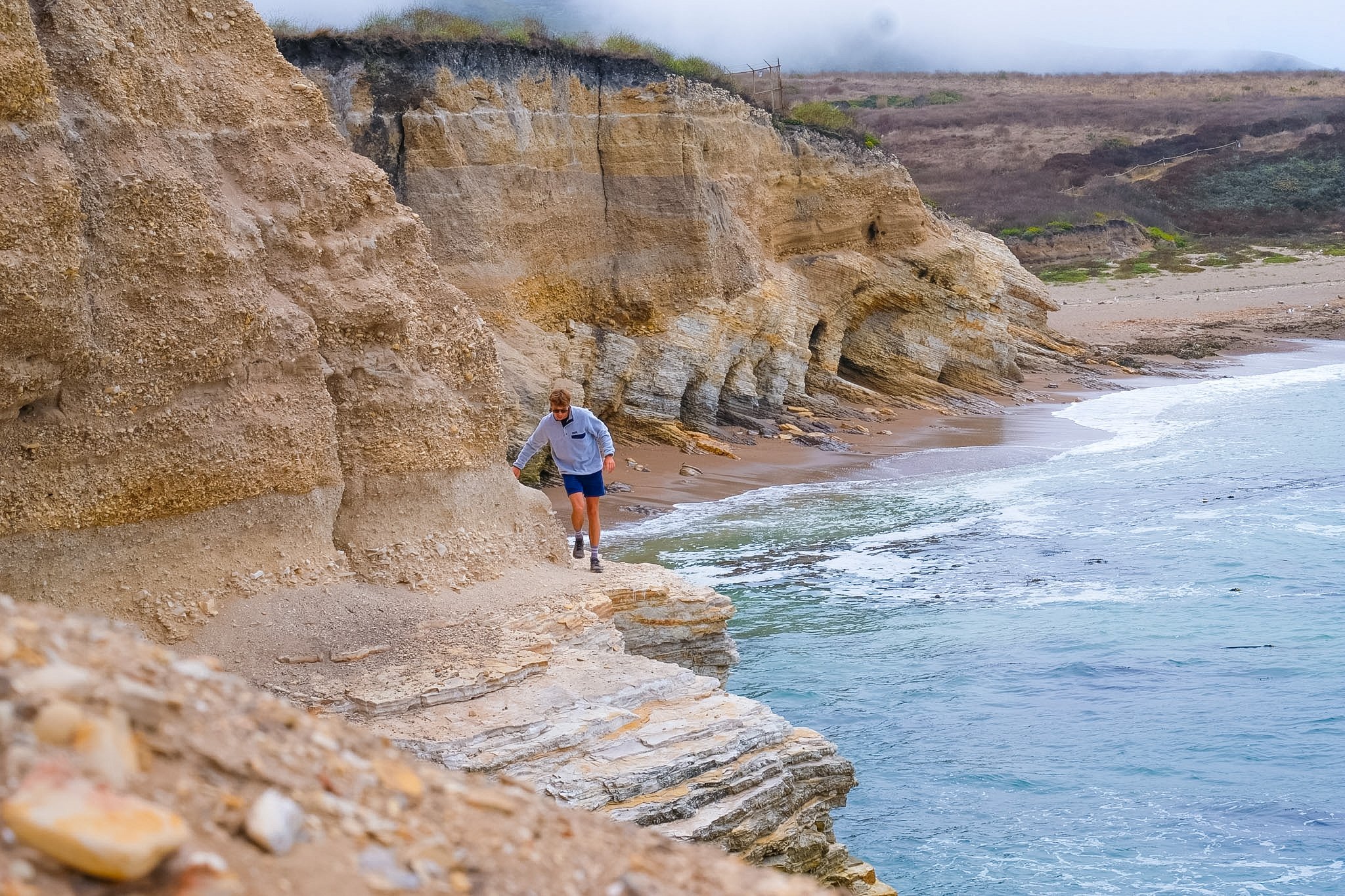 A person in a white jacket and shorts walking along a sandy and rocky beach near large beige and layered cliffs, with the ocean on the right and a cloudy sky above.