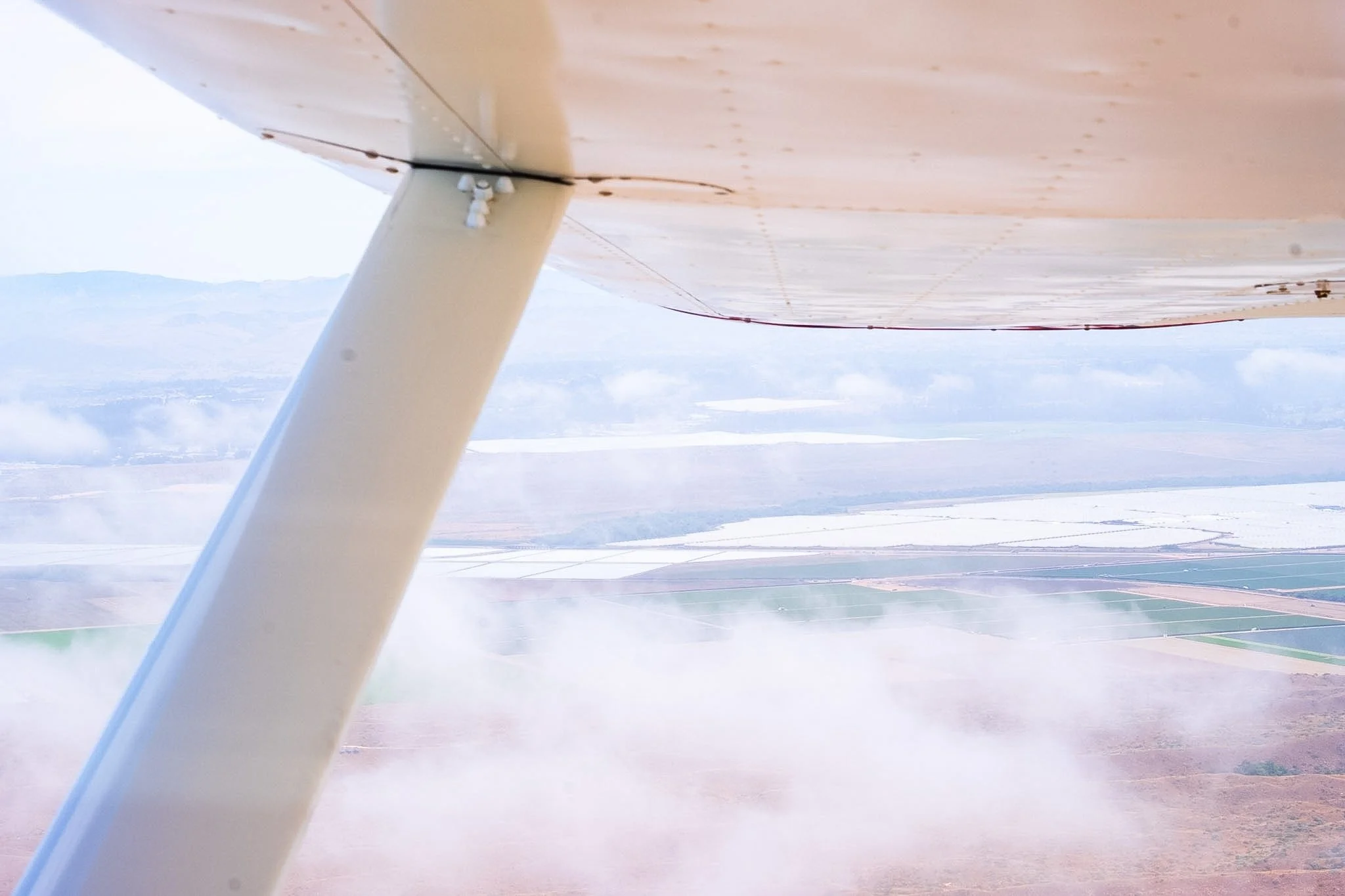 Aerial view from a small aircraft showing fields and a landscape below, with part of the plane's wing and support strut visible in the foreground.