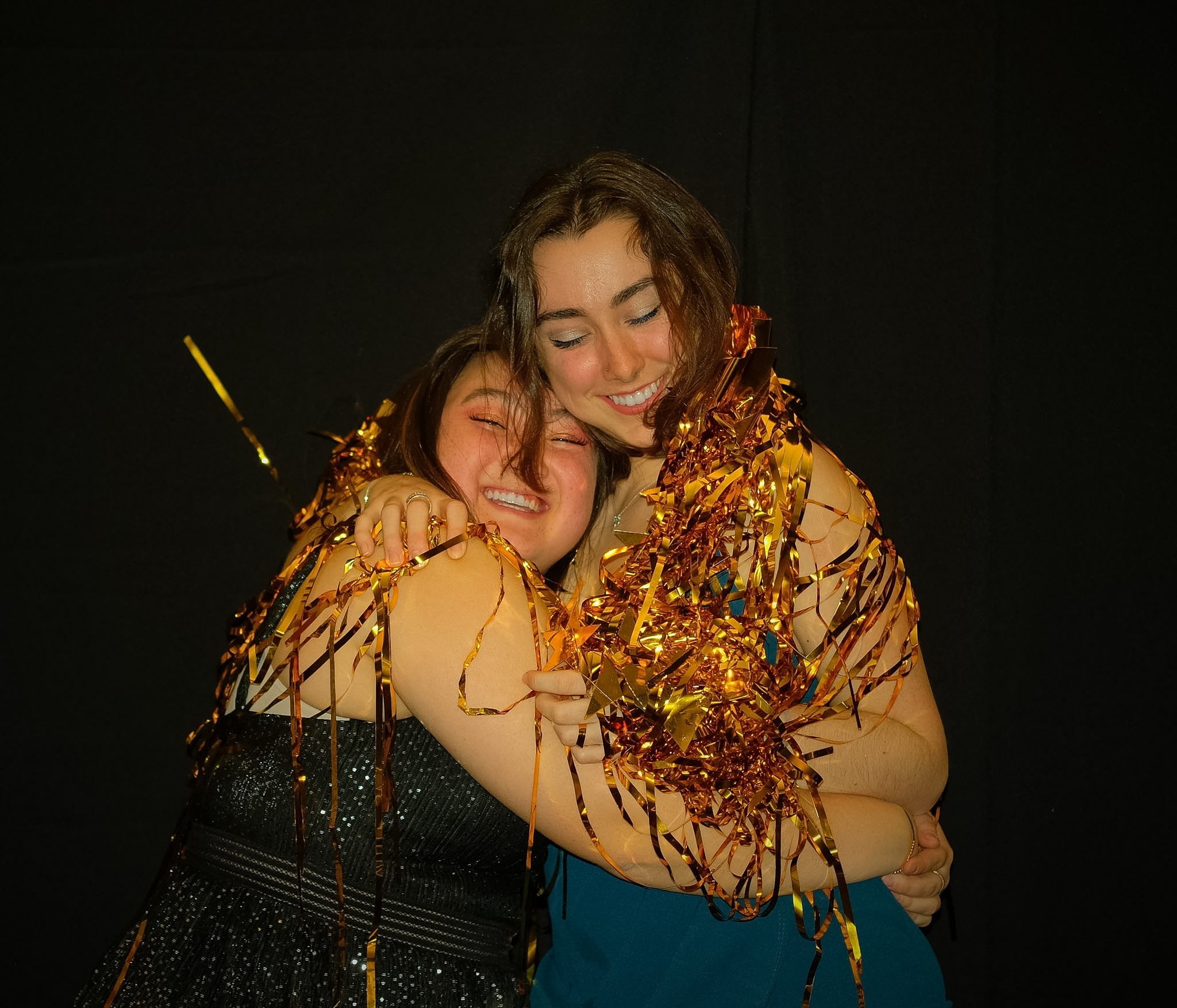 Two women hugging each other tightly, smiling with eyes closed, wrapped in shiny gold and orange confetti, against a black background.
