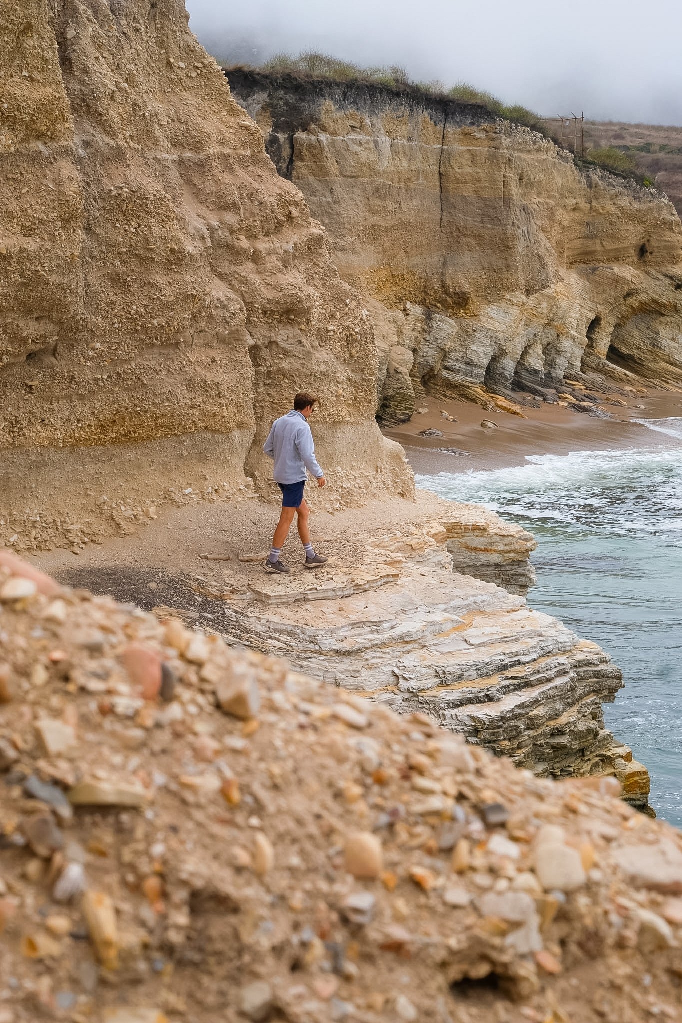 A person walking along a rocky beach with large cliffs in the background and fog in the sky.