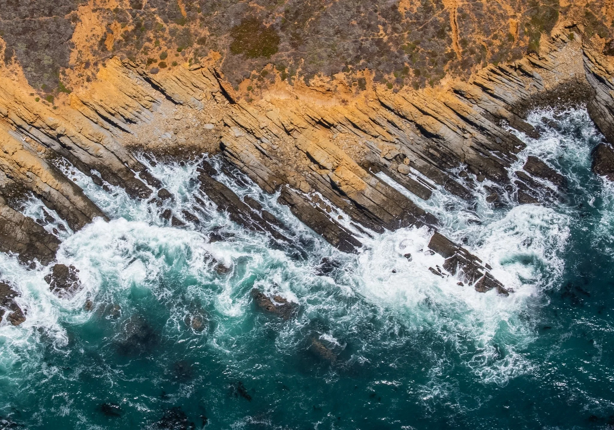Aerial view of rugged coastline with orange-brown cliffs and rough turquoise ocean waves crashing against rocks.