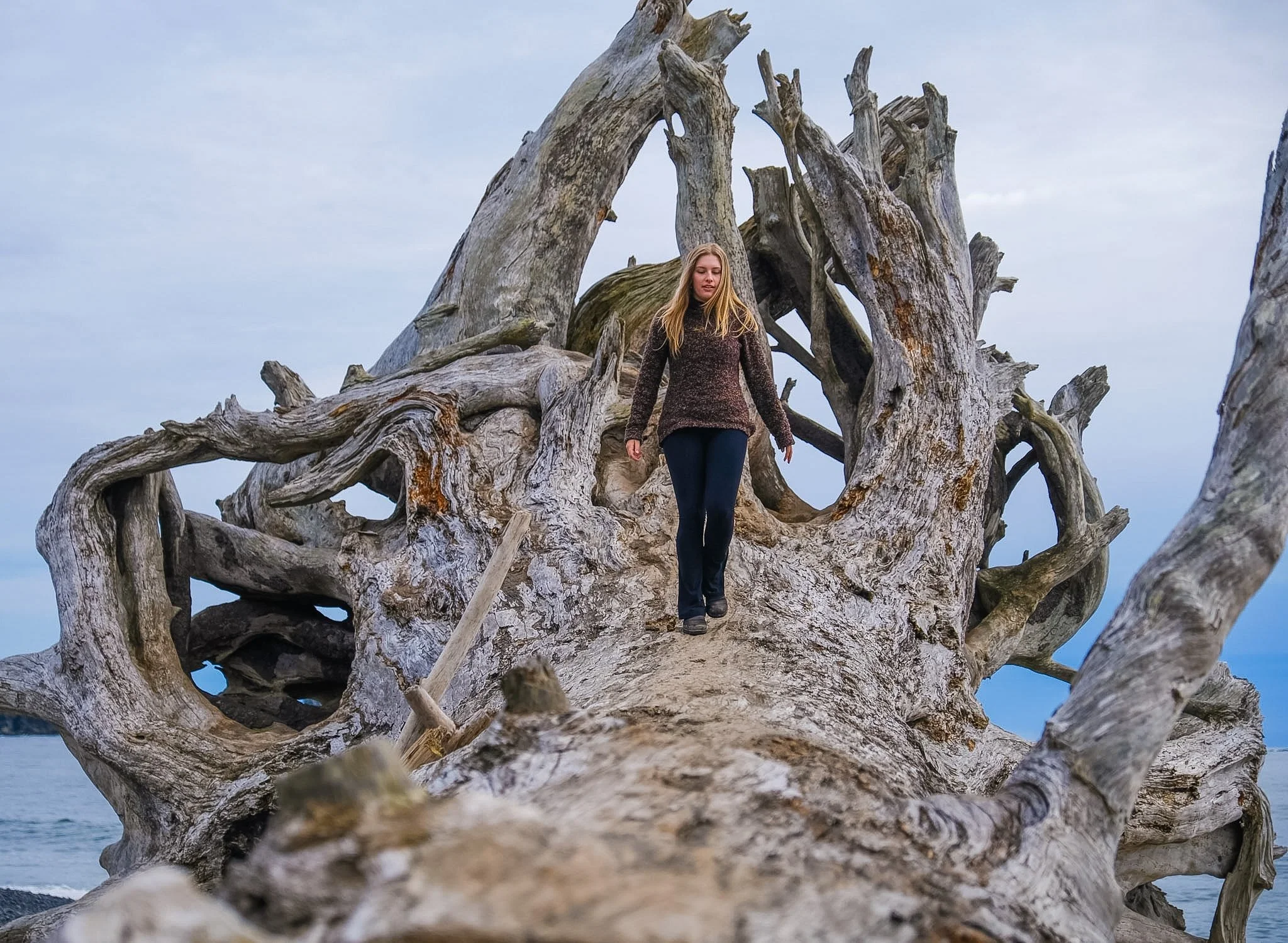 A young woman with blonde hair wearing a long-sleeve shirt and dark pants walking on large driftwood at the beach during overcast weather.