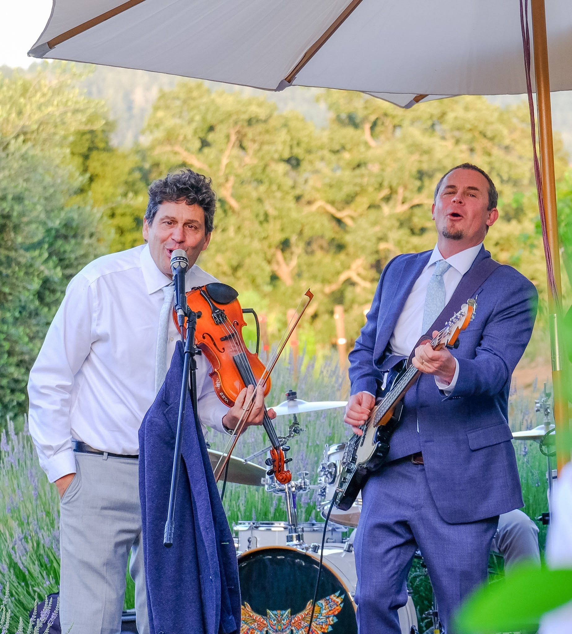 Two men dressed in formal attire, one playing a violin and the other playing a guitar, under a large umbrella outdoors with a background of green trees and grass.
