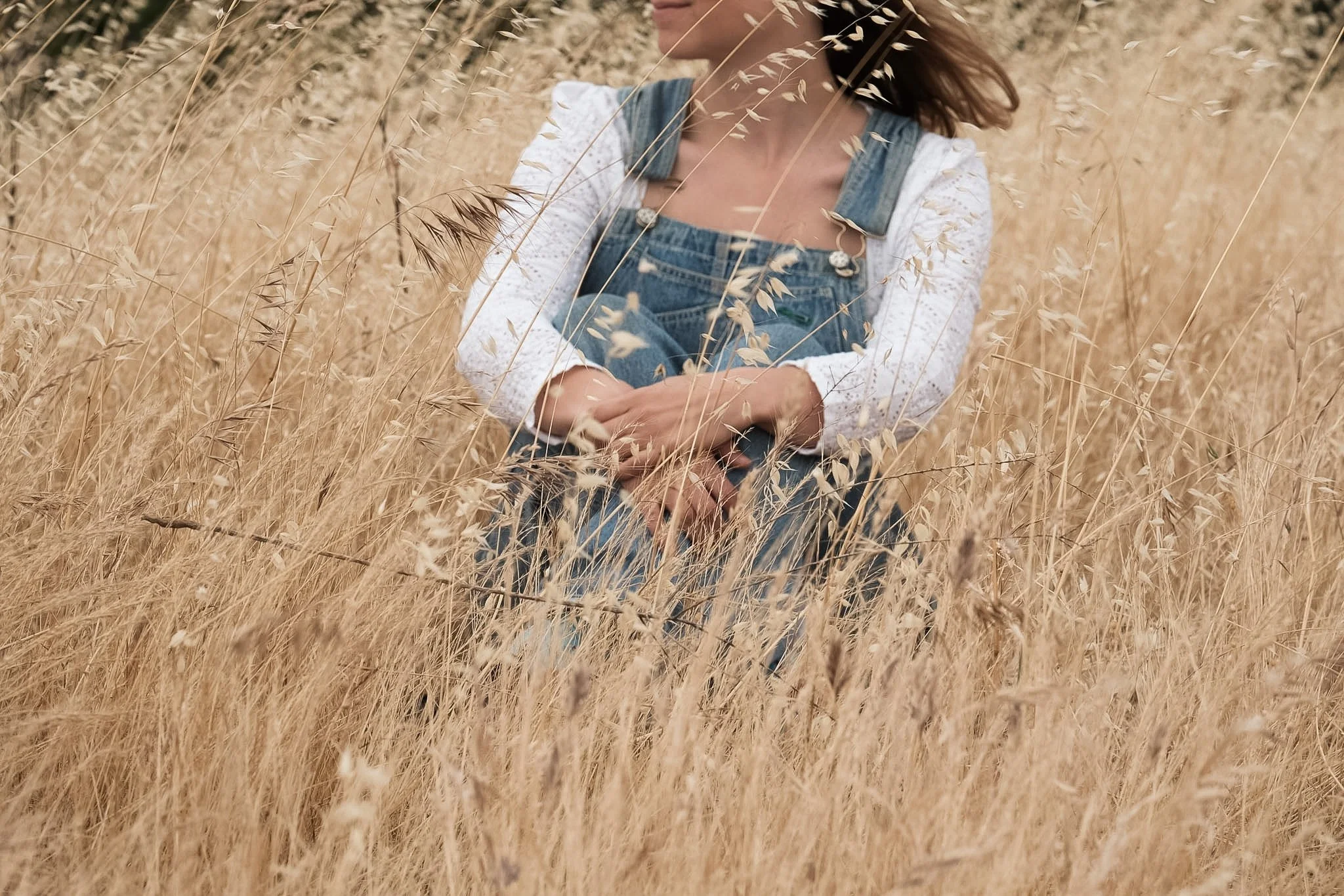 Woman in denim overalls and a white lace shirt sitting in a wheat field.