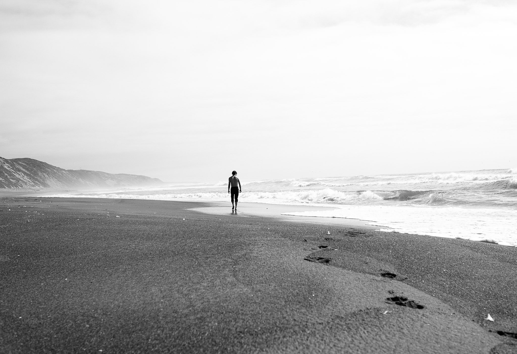 A person walking alone along the shoreline of a beach with waves, cliffs in the distance, and a cloudy sky.