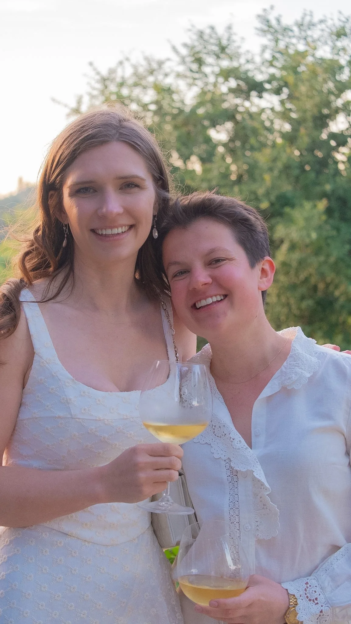 Two women smiling outdoors holding white wine glasses, wearing white dresses, with lush greenery and trees in the background during daylight.