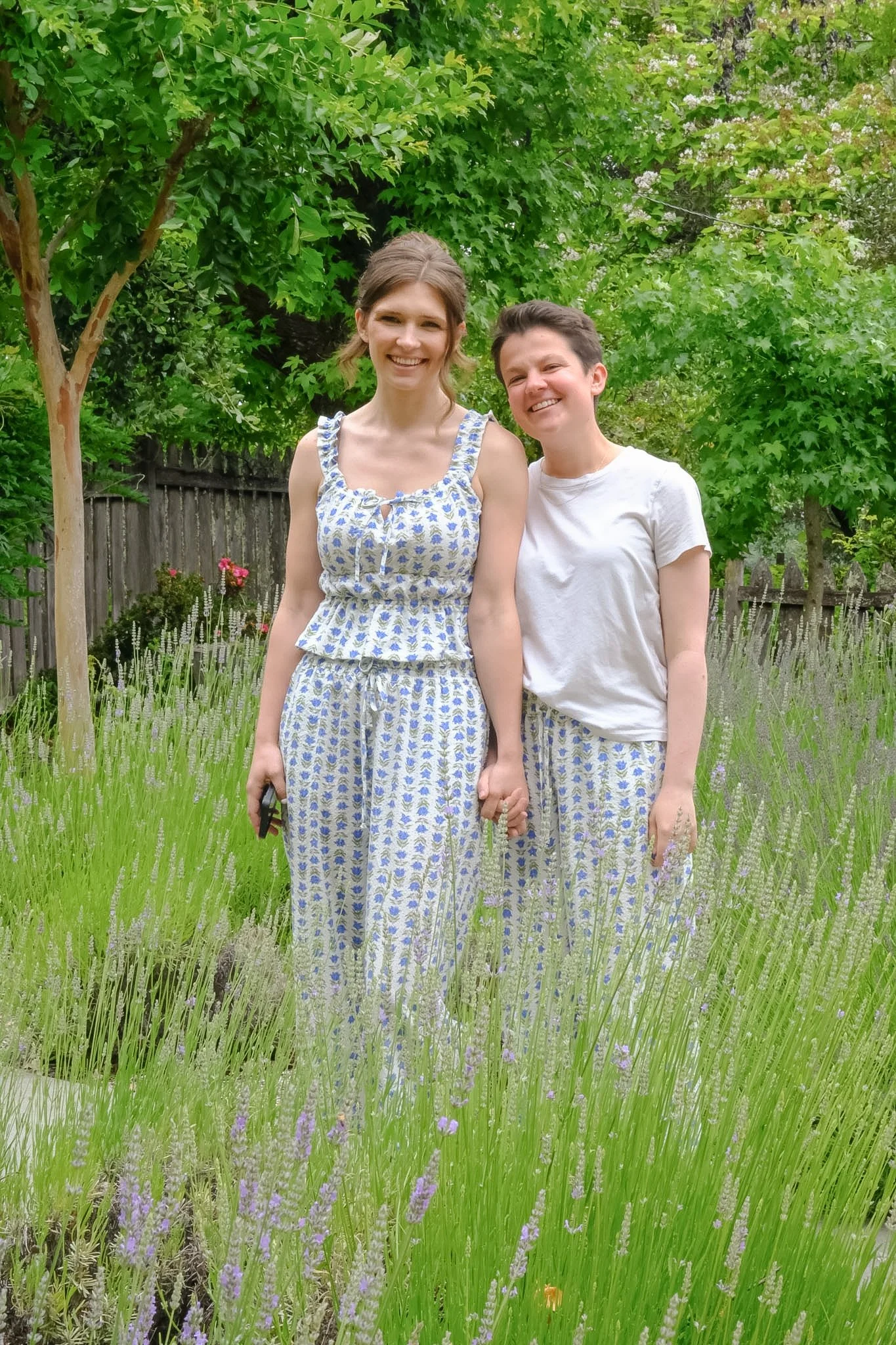 Two women smiling in a garden with green trees, purple flowers, and a wooden fence.