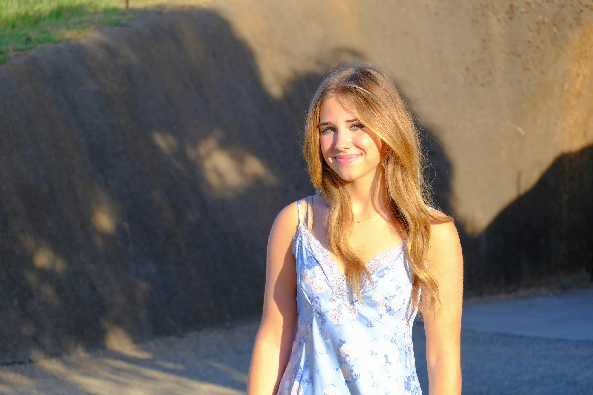 A young woman with long, wavy, blonde hair and blue eyes standing outdoors at sunset, wearing a white and blue floral dress, smiling gently at the camera.