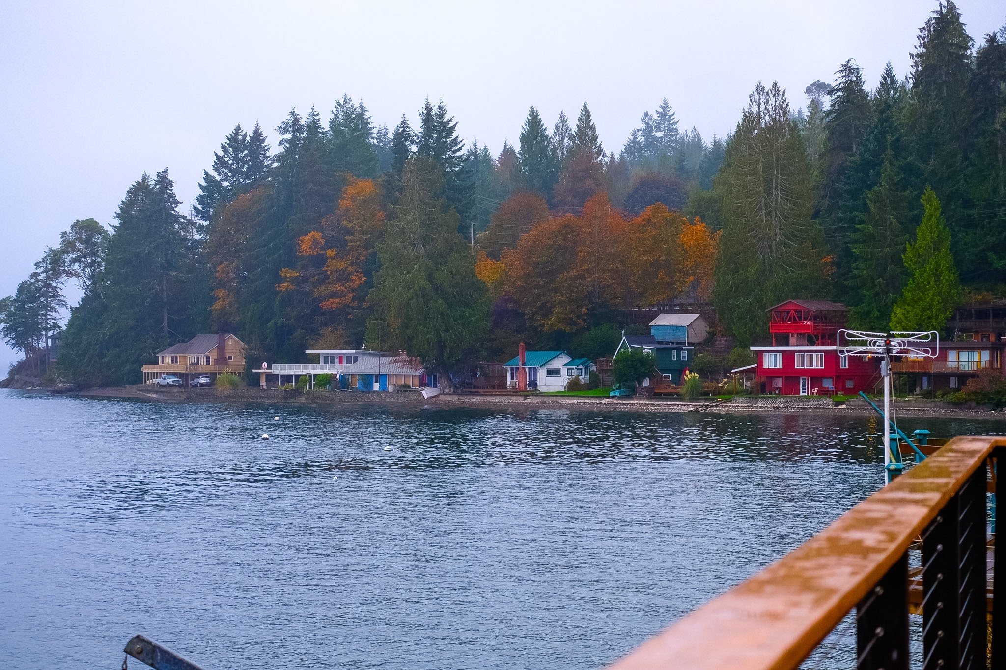 A lakeside scene with colorful houses at the water's edge, trees with fall foliage on a foggy day.