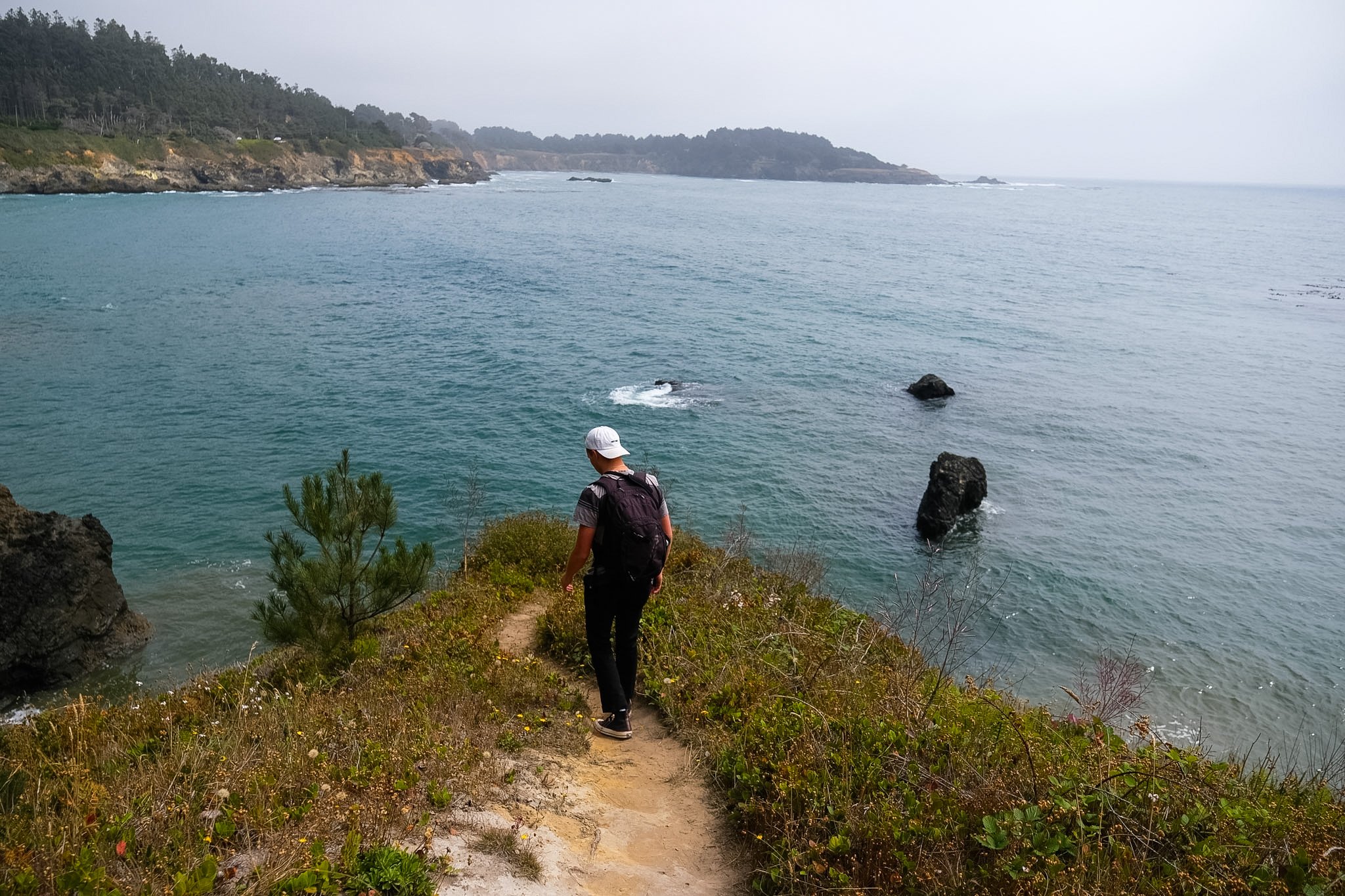 A person walking on a dirt trail along the coast with water and rocky cliffs in the background, overcast sky.
