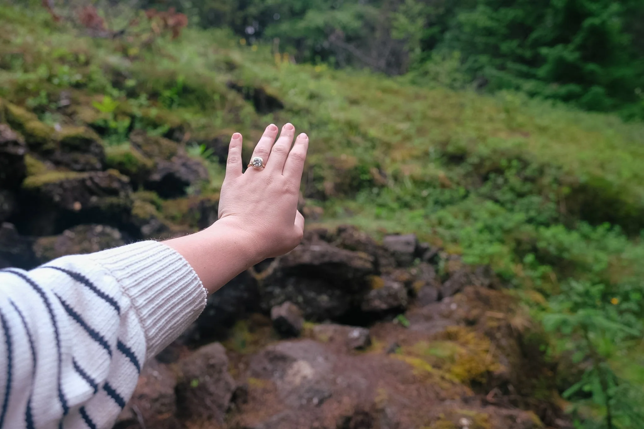 A person's hand with a wedding ring reaching out to a lush green forest and rocky stream in the background.