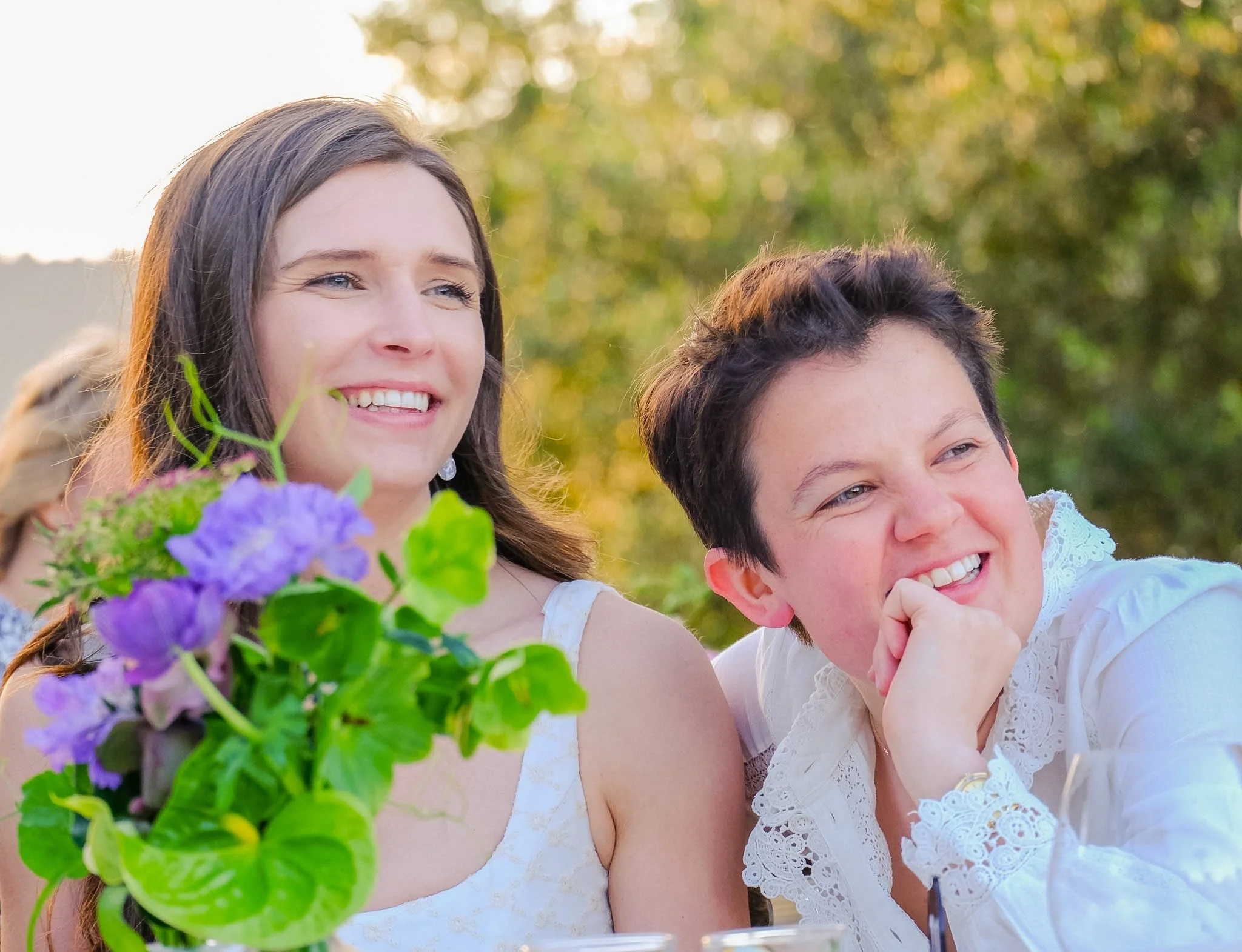 Smiling young woman and young man enjoying outdoors with greenery in background, person holding purple flowers in foreground, both wearing light-colored clothing.
