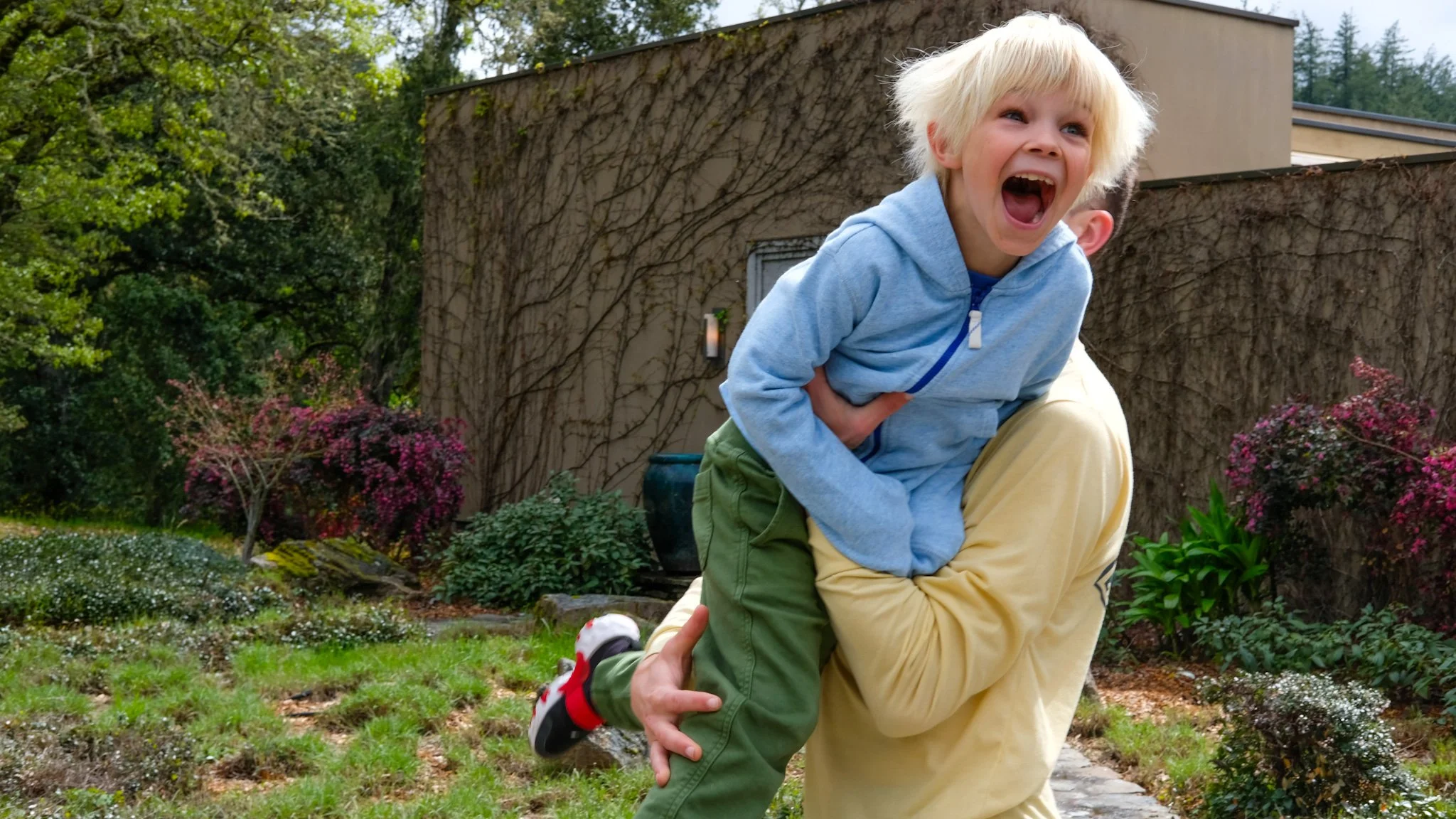 A young child with blonde hair and an open-mouthed smile is being lifted by an adult in a garden with trees, bushes, and colorful flowers.
