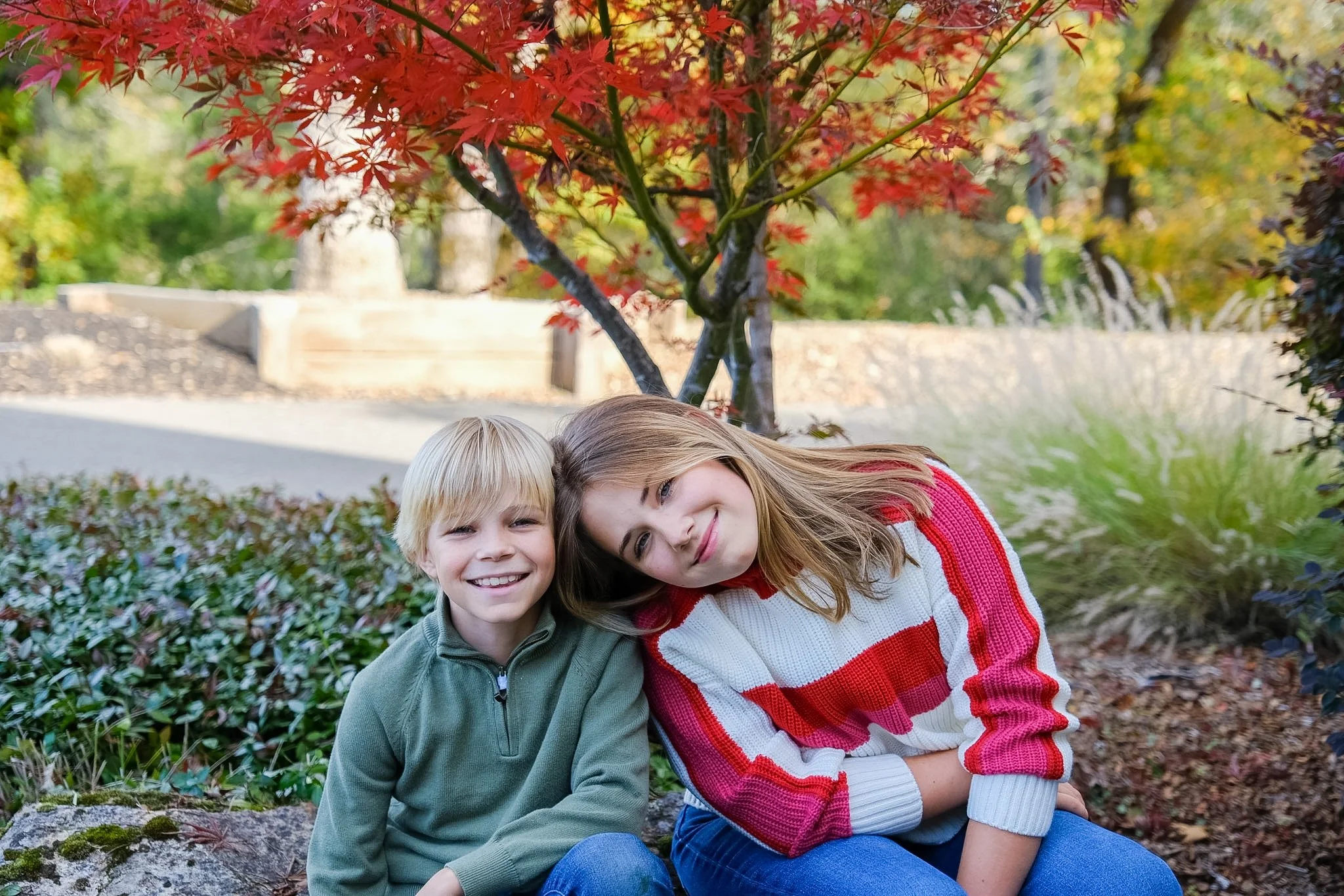 Two children, a boy and a girl, sitting close together outdoors under a red maple tree, smiling at the camera.