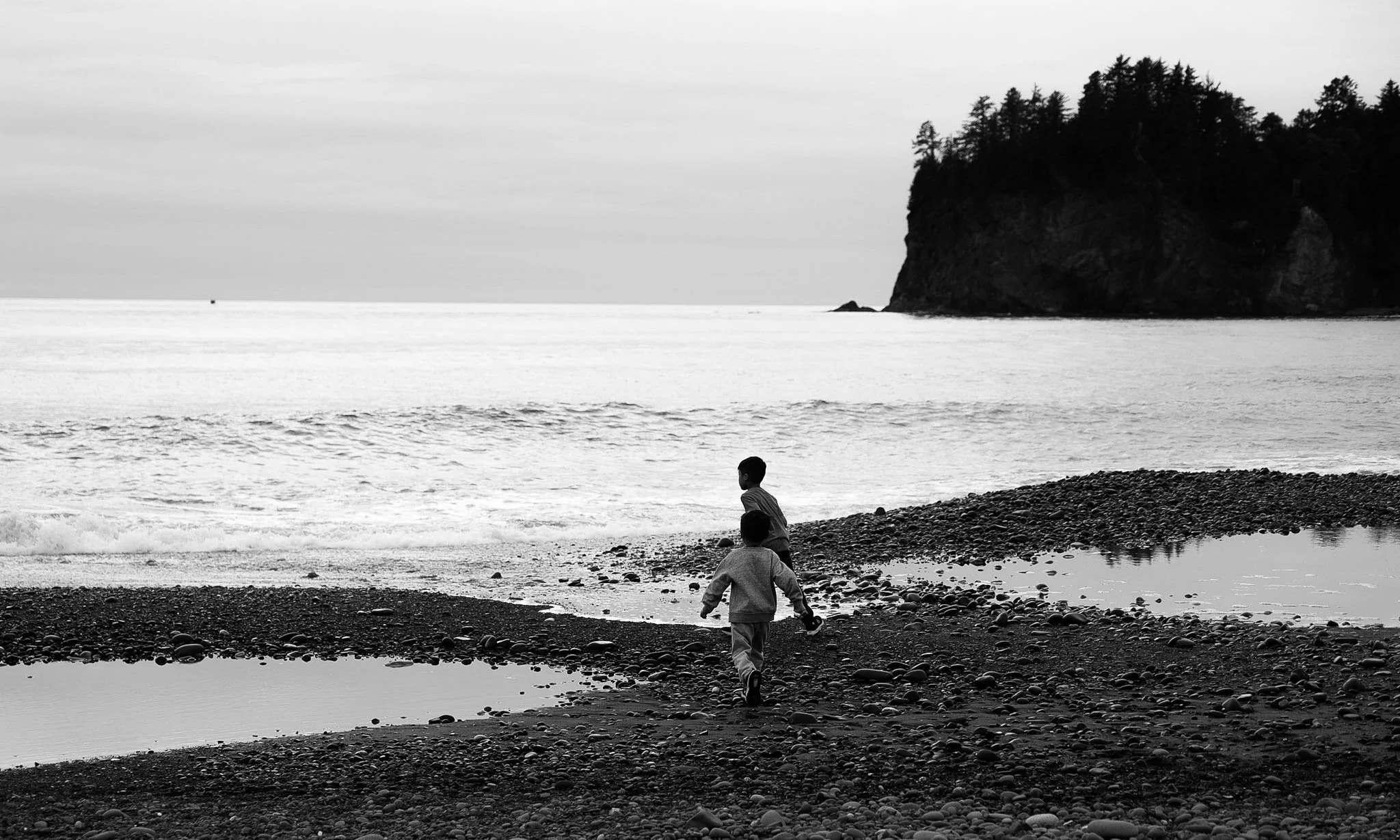 Black and white photo of two children on a pebble-covered beach near the water, with a large rock formation and trees in the background.