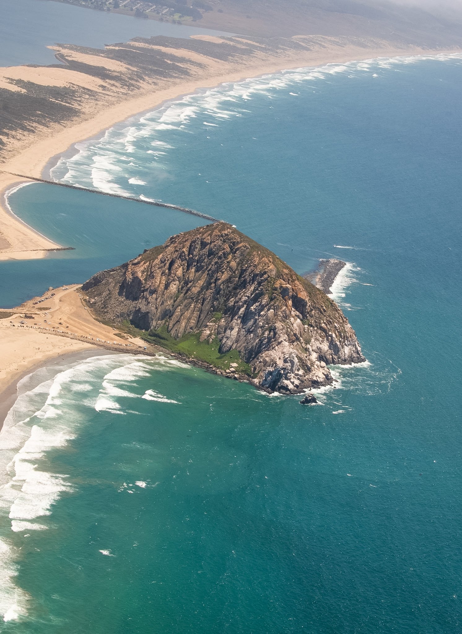 Aerial view of a stretch of beach with waves crashing on the shore, a rocky cliff, and a saltwater lagoon separated from the ocean by a sandbar, with land and water in the background.