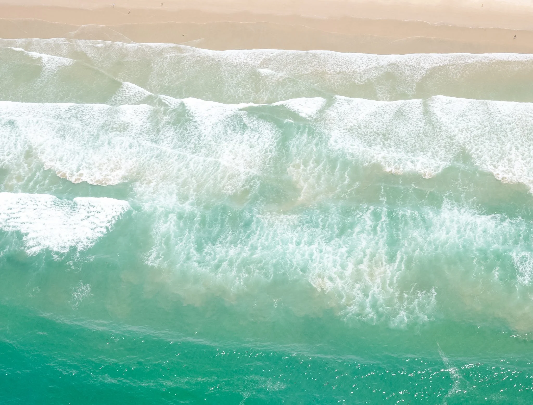 Aerial view of ocean waves crashing onto sandy beach with some boats in the distance.