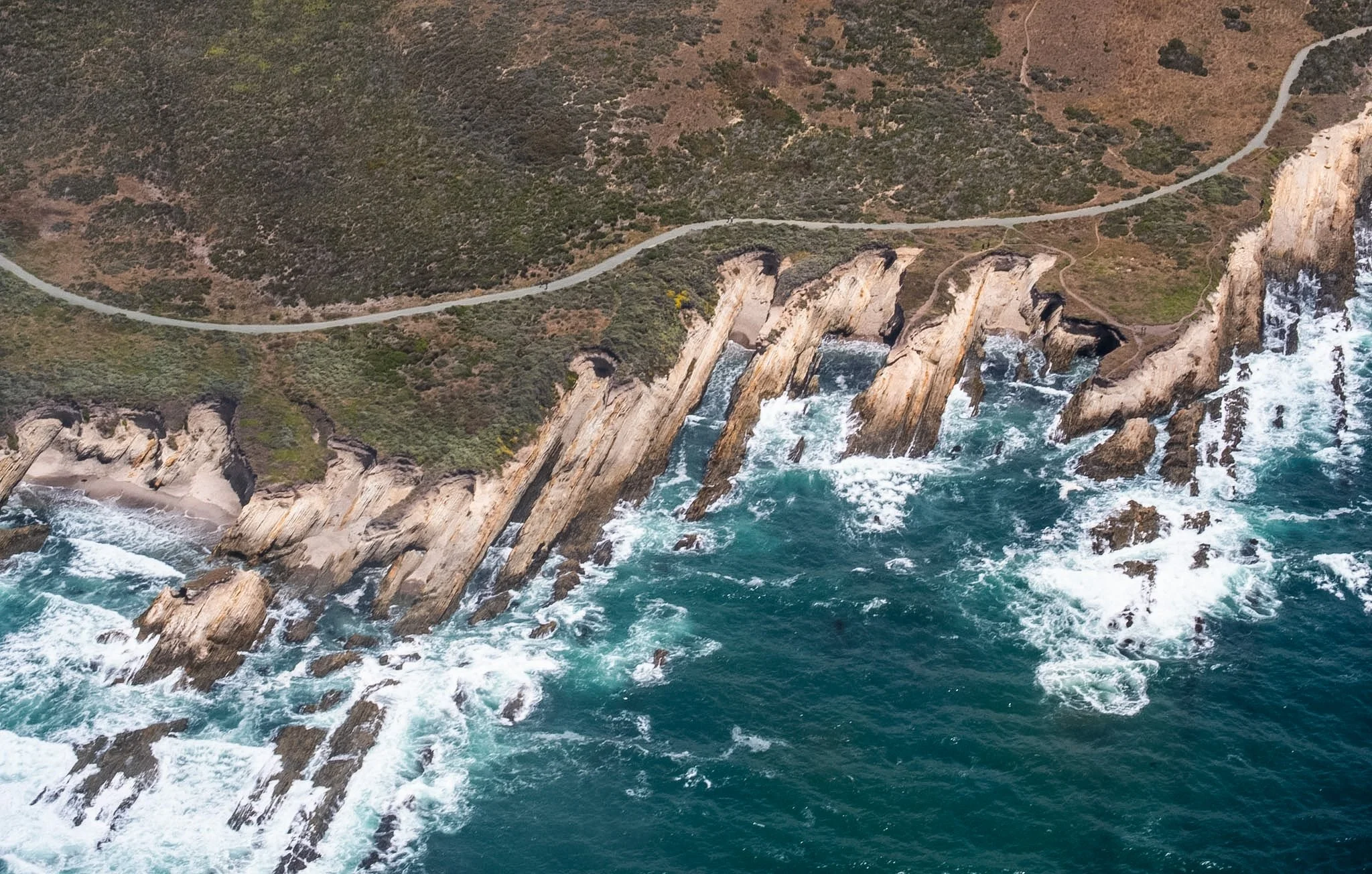 Aerial view of a coastal landscape with rugged cliffs, a winding road, and rough ocean waves crashing against rocks.
