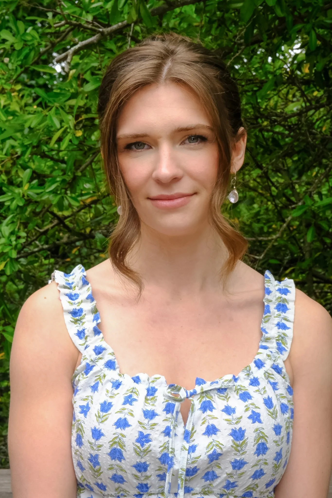 A woman with shoulder-length brown hair and blue eyes, wearing a white dress with blue floral pattern and earrings, standing in front of green foliage.