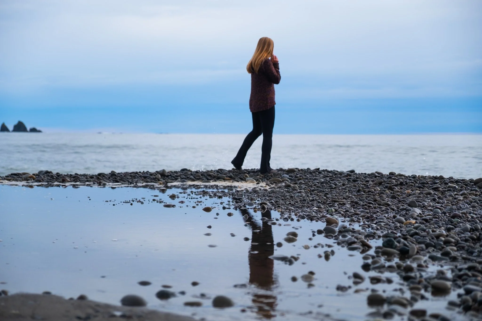 A woman standing on a pebbled beach looking at the water, with mountainous formations in the distance and a cloudy sky overhead.