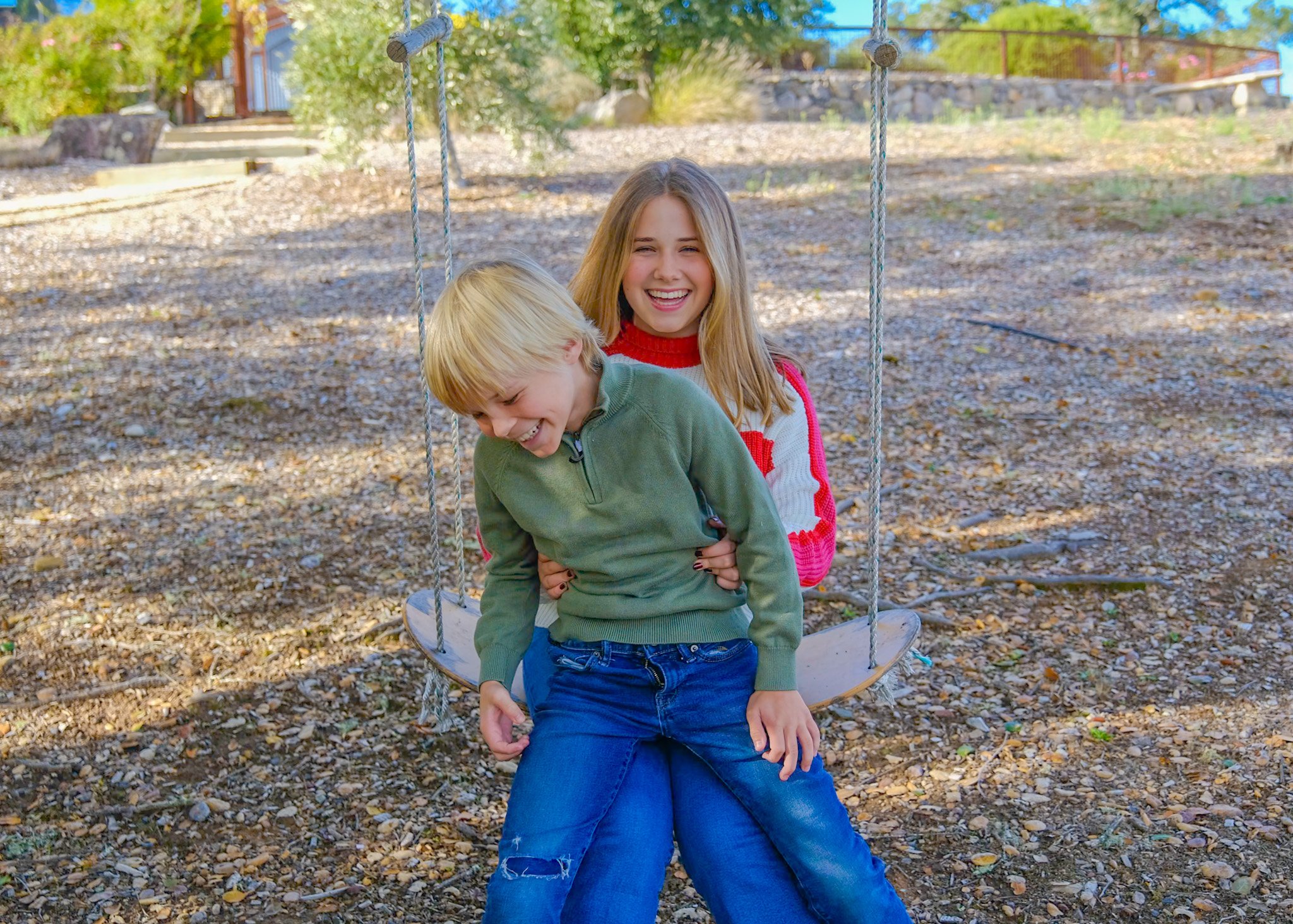 Two children, a girl and a boy, are sitting on a swing outdoors, surrounded by fallen autumn leaves. The girl is smiling and wearing a red and white sweater, while the boy is looking down and wearing a green sweater. The background features trees, a stone wall, and a backyard area.