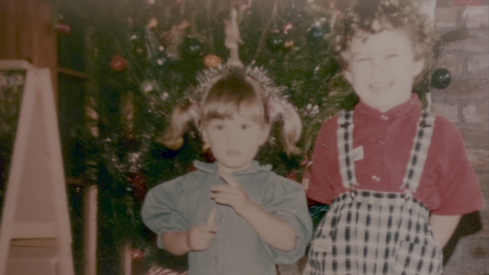 Two children standing in front of a decorated Christmas tree, with one girl with pigtails and a boy with glasses and floppy hair smiling.