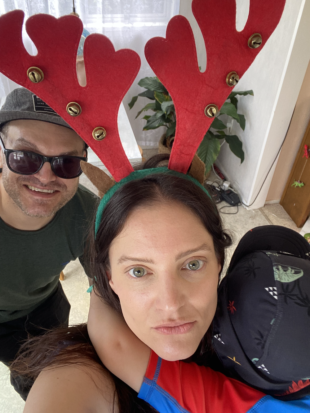 Two people celebrating Christmas, with the woman wearing red reindeer antlers and the man wearing sunglasses and a hat.