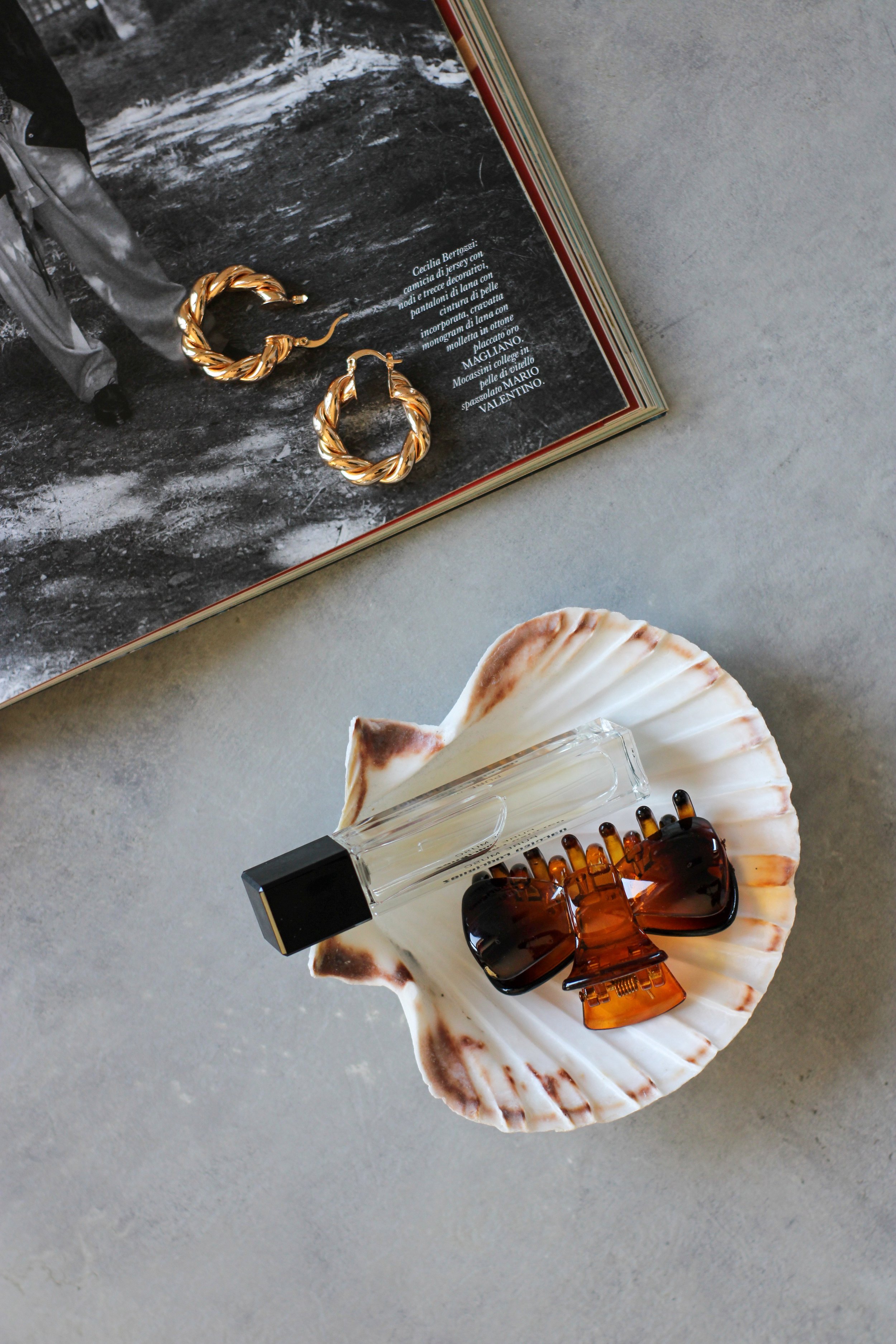 Gold twisted hoop earrings, a seashell, a glass perfume bottle, and a brown hair clip with a tortoise shell pattern on a large seashell, all placed on a marble surface.