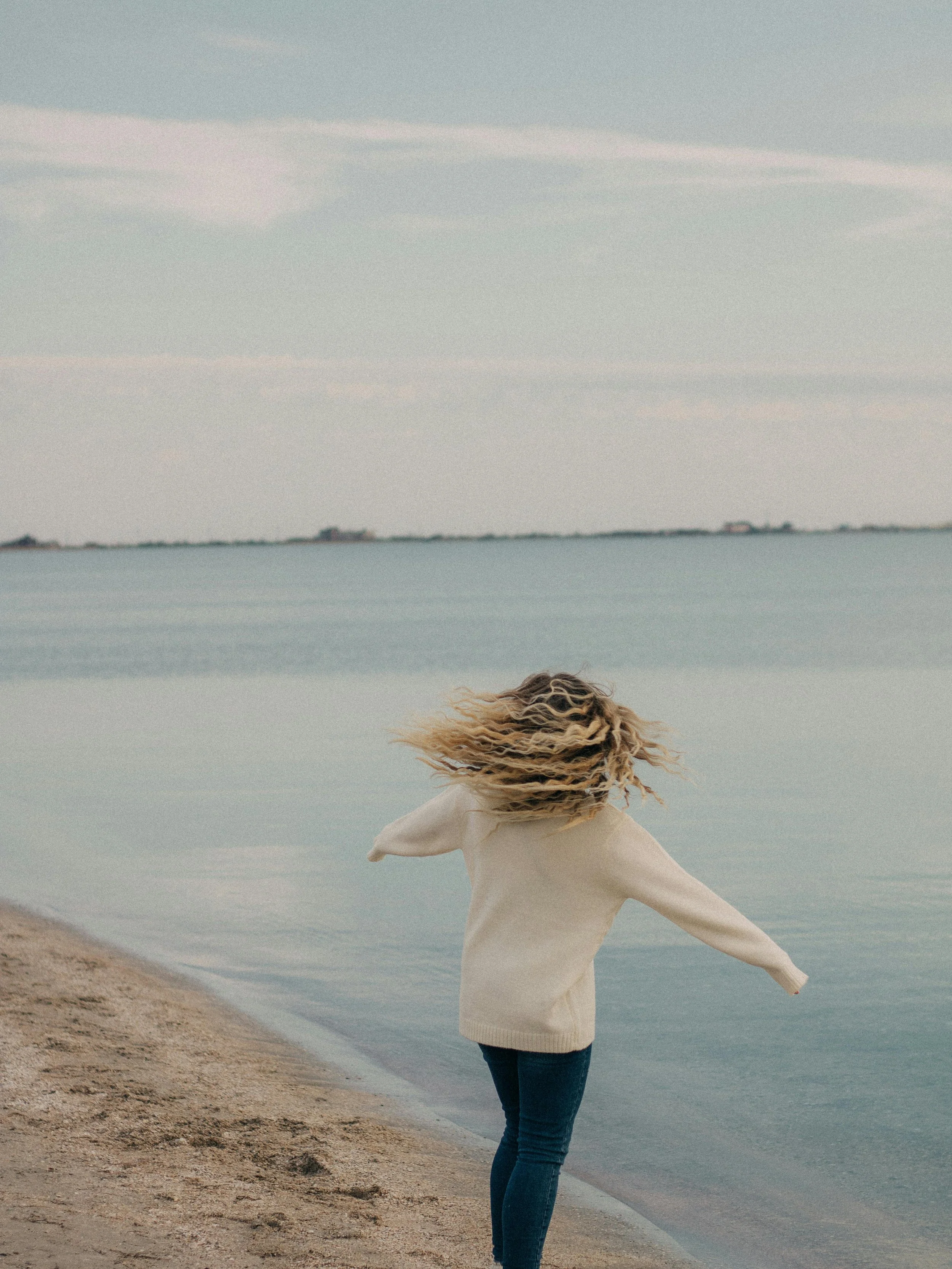 A person in a beige sweater and jeans standing on a sandy beach with arms outstretched, facing the water, with hair blowing in the wind, and a distant shoreline visible across the water.