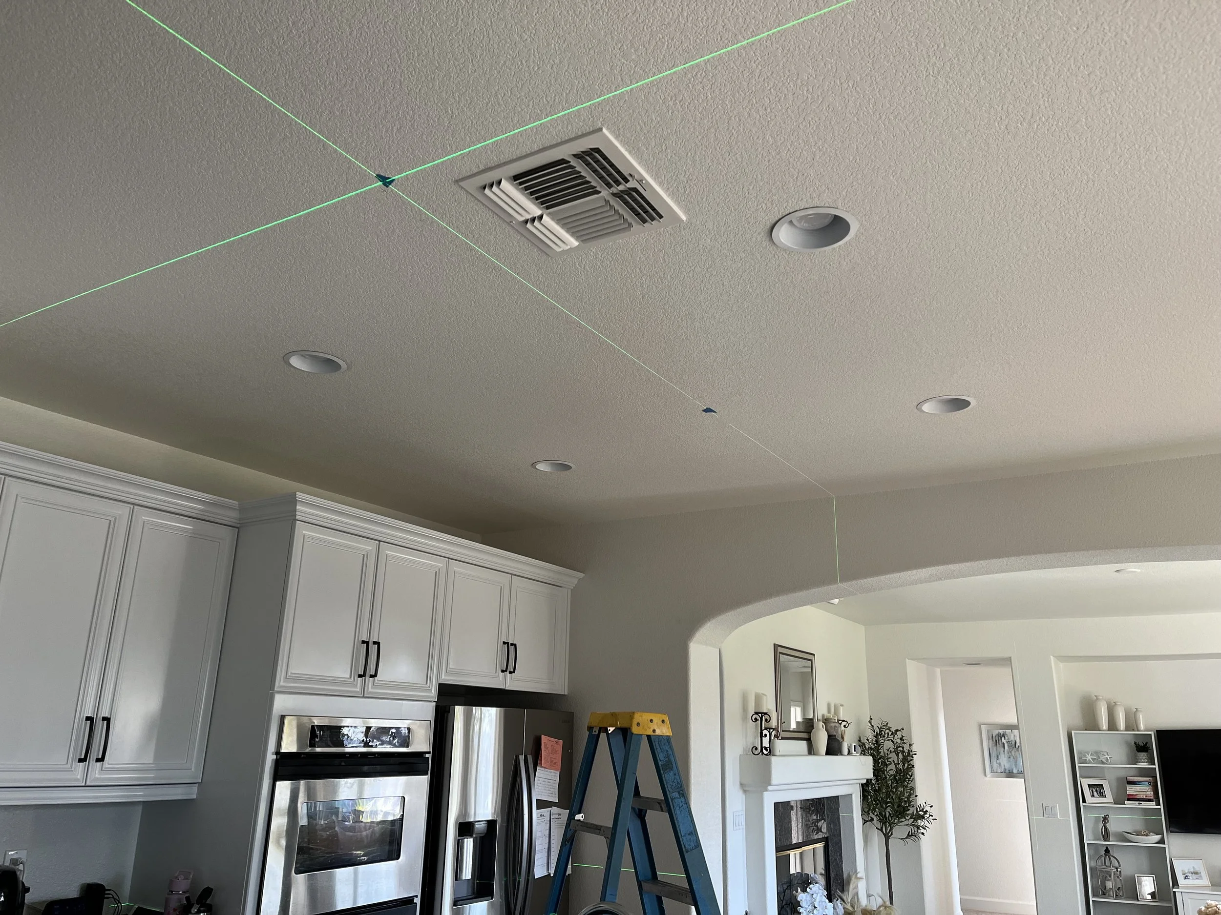 Ceiling with air vent, recessed lights, and laser line projection in a kitchen with white cabinetry and a living area in the background.