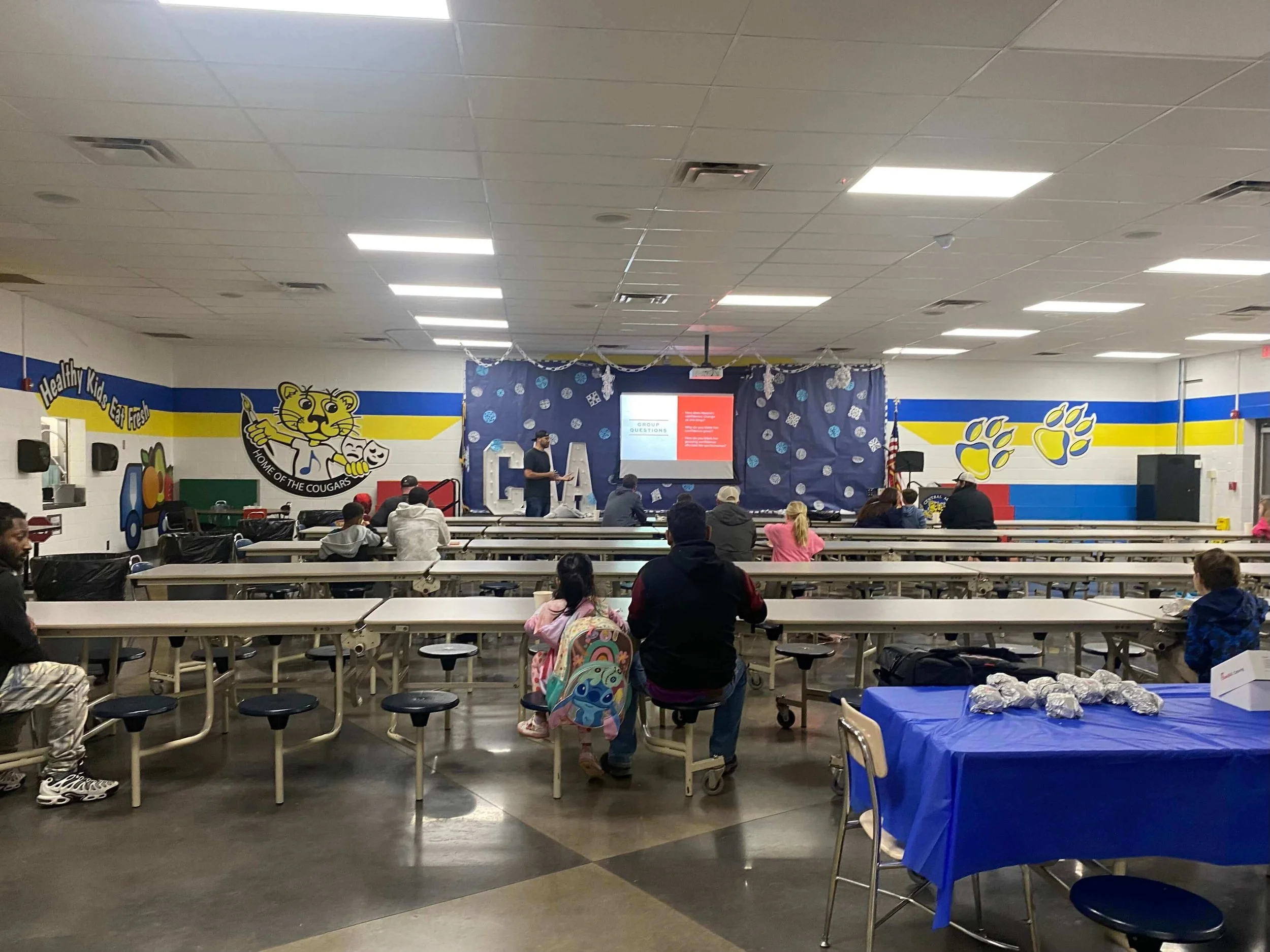 A school cafeteria with students sitting at tables, a stage decorated with blue curtains and winter-themed decorations, and a large mascot graphic on the wall promoting the school as the home of the Cougars.