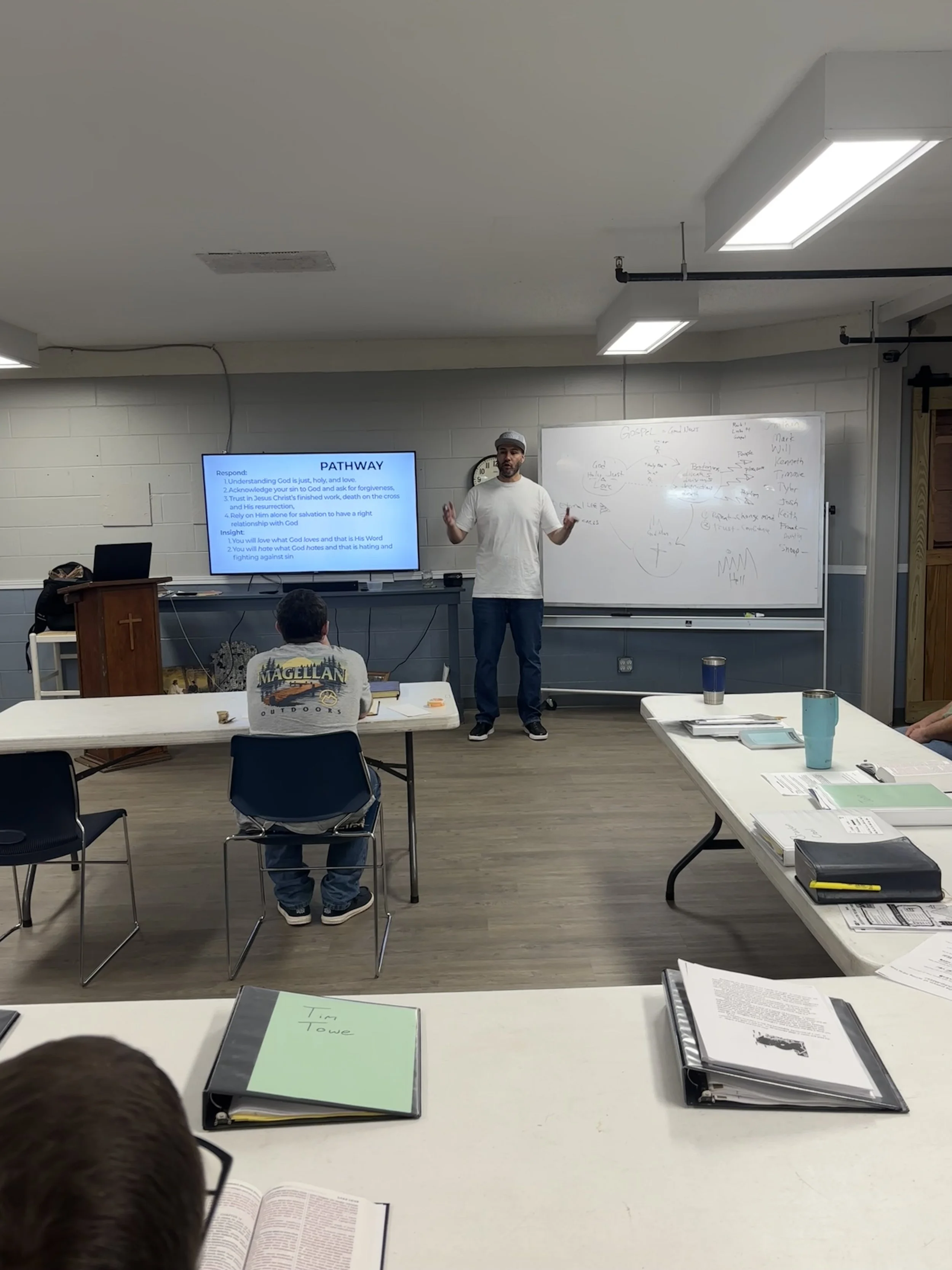 A classroom lecture with a man in a white t-shirt and cap standing in front of a whiteboard and a screen displaying a presentation titled 'PATHWAY'. Students are seated at tables with notebooks, folders, and drinks, listening and taking notes.