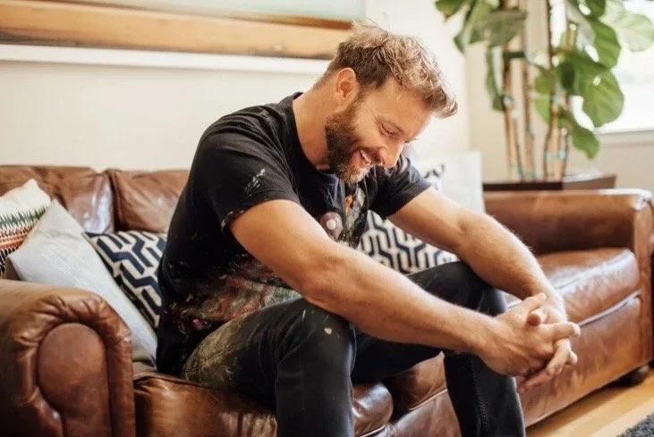 A man with a beard and curly hair sitting on a brown leather couch, smiling and laughing with his hands clasped, in a cozy living room with plants and decorative pillows.