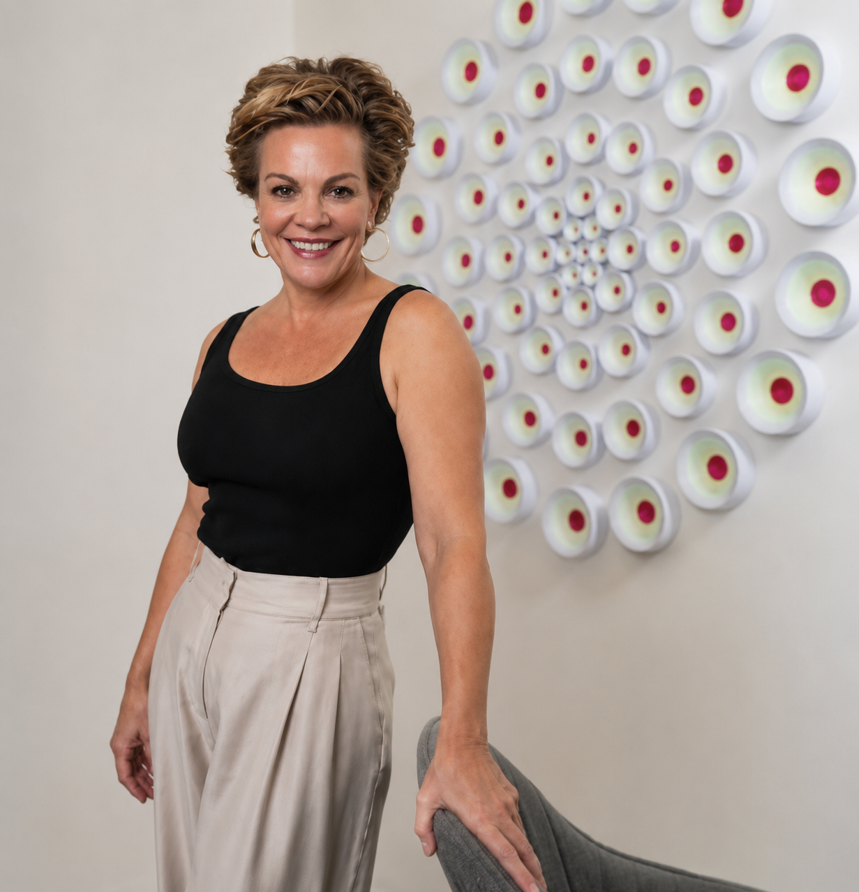 A smiling woman with short, curly hair wearing a black sleeveless top and beige pants, standing in front of a wall with circular wall art.