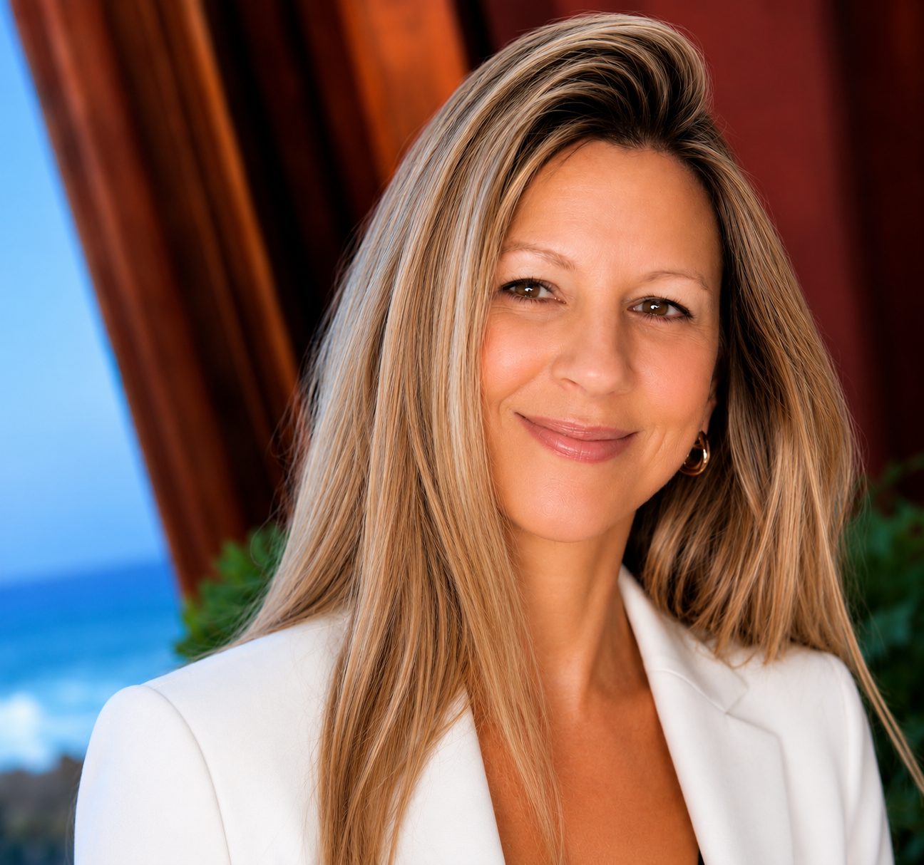 A woman with long, blonde hair smiling at the camera, dressed in a white blazer, with wooden structures and a coast with blue water.