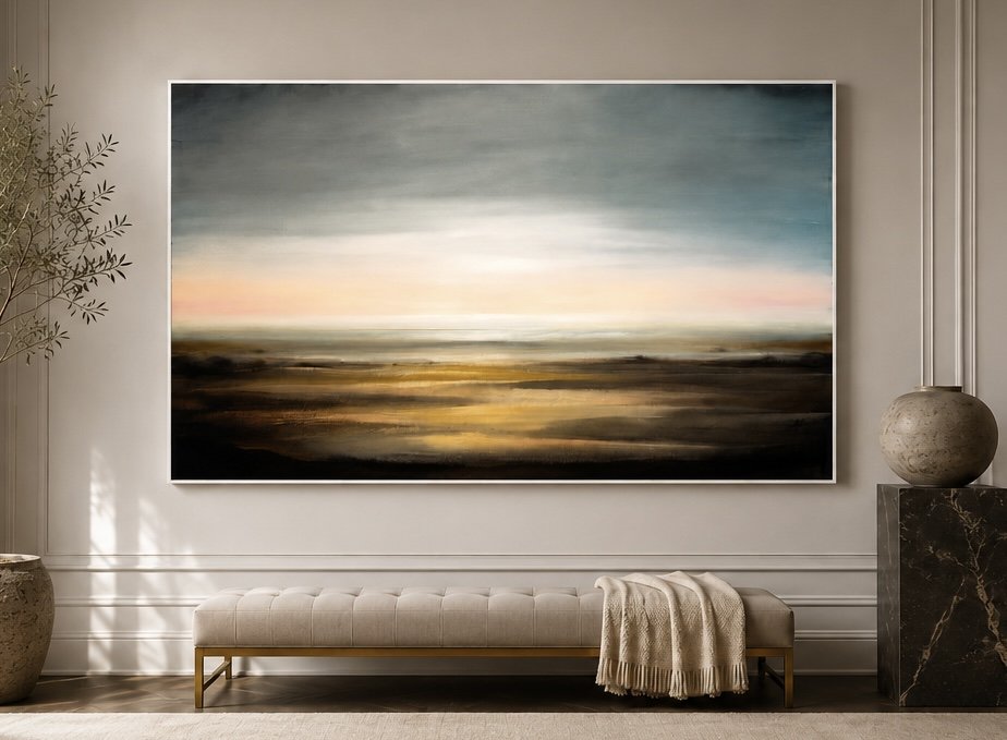 An abstract landscape painting with soft pastel colors hangs on a white wall in a modern room. The room features a beige tufted bench, a cream throw blanket, a large light-colored vases on the left and right, and minimal decor.