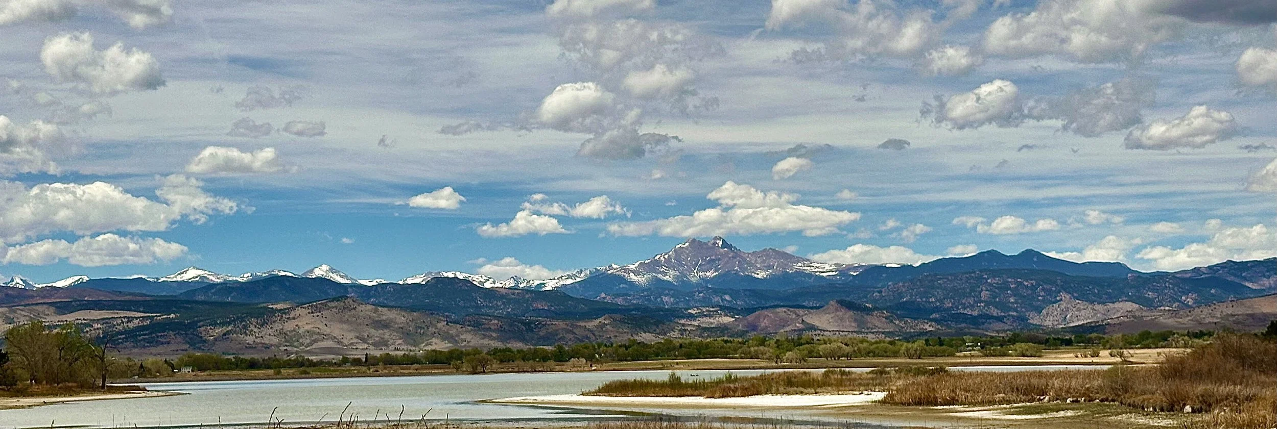 Scenic view of distant snowy mountain range with partly cloudy sky above, a body of water in the foreground, and some trees and shrubs along the shoreline.