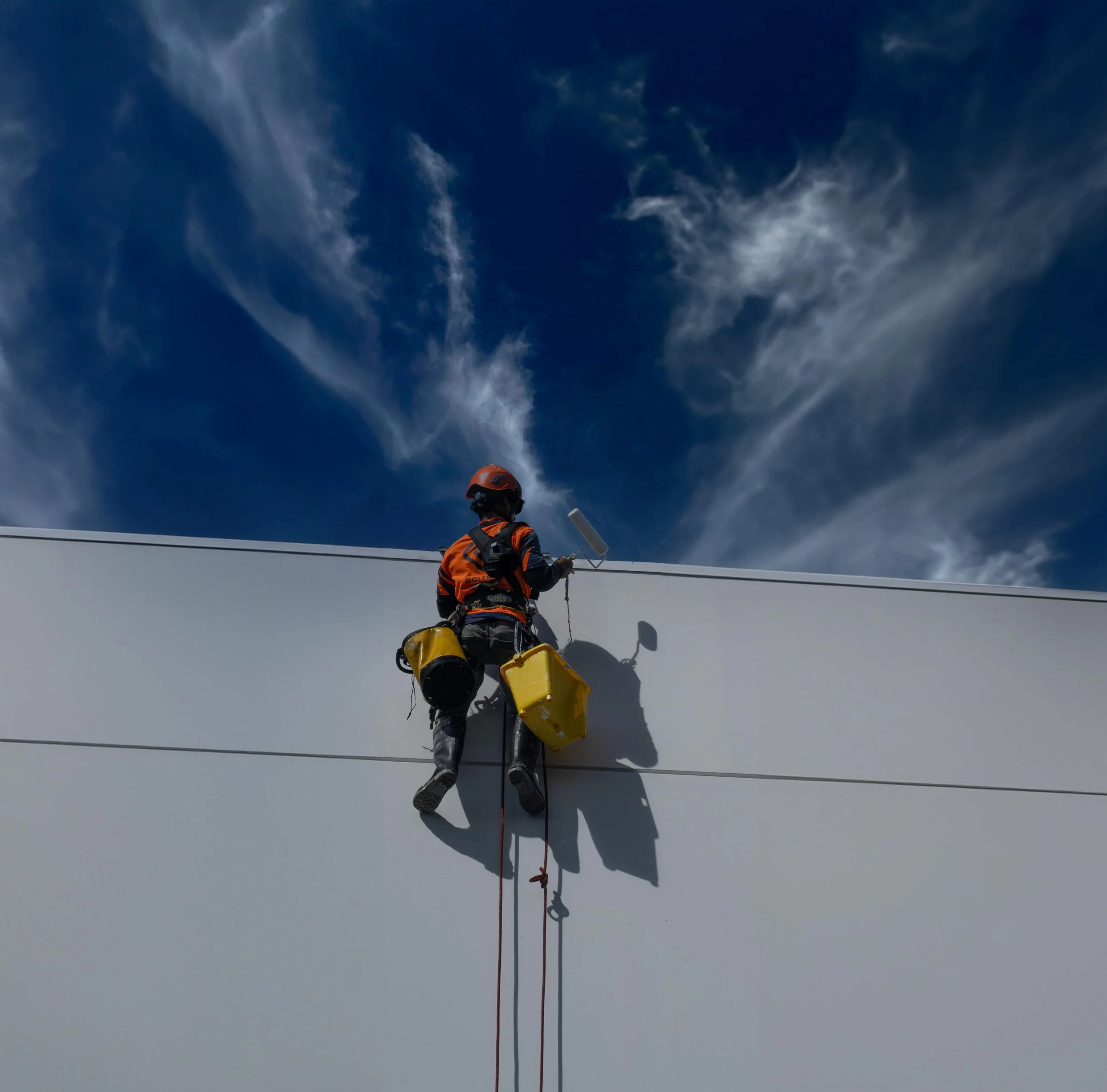 A person in outdoor gear, including a helmet and harness, appears to be climbing or rappelling on a snow or ice-covered surface, with a view of a cloudy sky above.