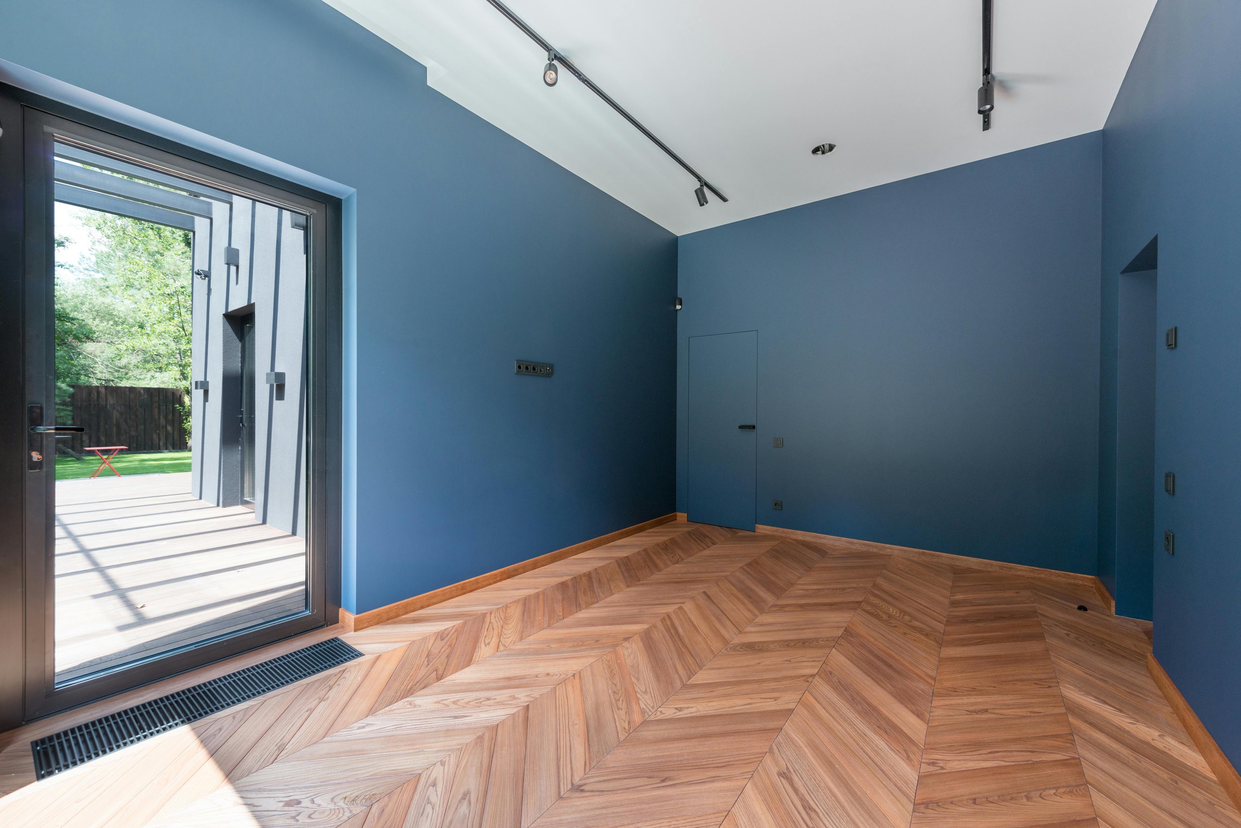 Interior of a room with blue walls and wooden herringbone floor, an open glass door leading outside, and track lighting on the ceiling.