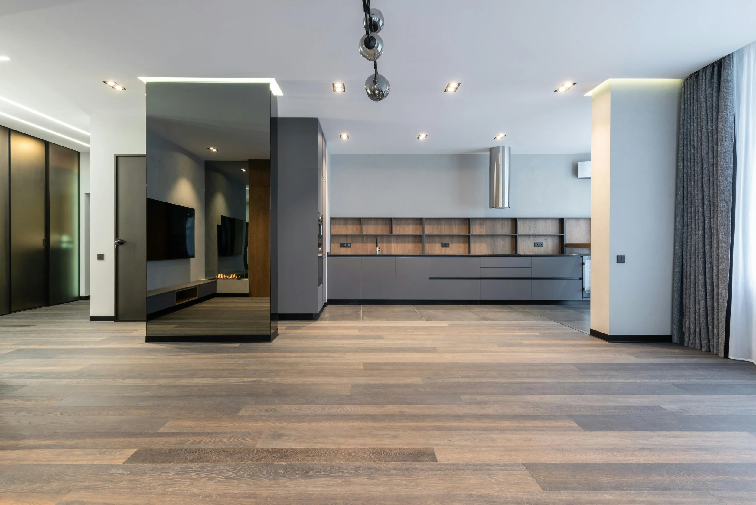 Modern kitchen with gray cabinets, a wood backsplash, and pendant lights, adjacent to a living area with a flat-screen TV and curtains.