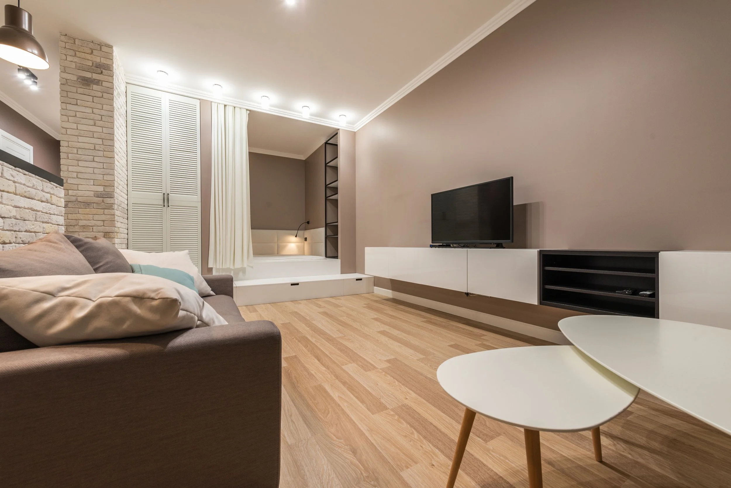 Living room with a gray sofa, beige pillows, a white coffee table, a wall-mounted TV, and a nook with white curtains and built-in shelves, hardwood flooring, and ceiling lights.