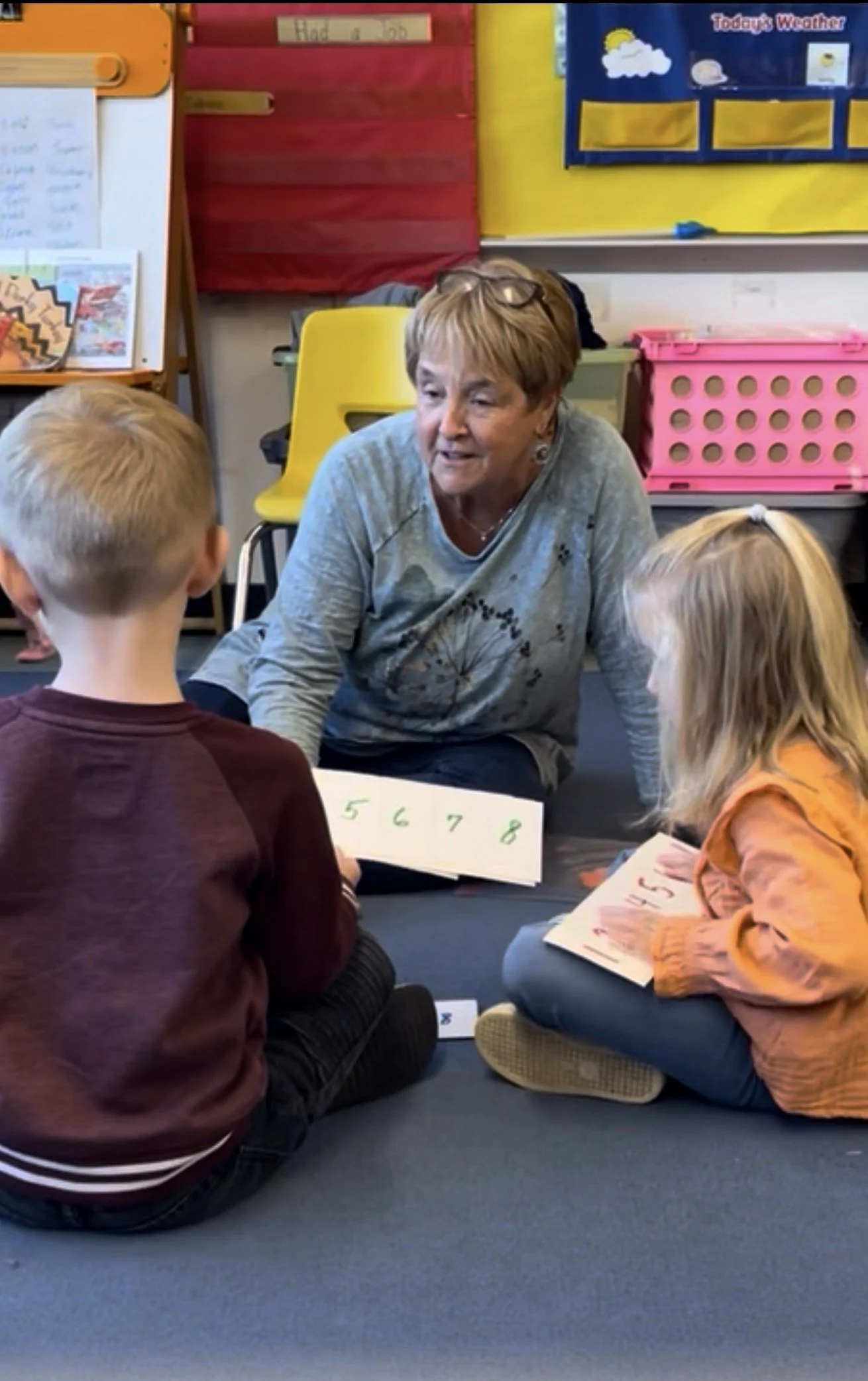 A woman is sitting on the floor with three children, engaged in a learning activity with number cards from 5 to 8. The classroom background includes colorful wall decorations and bins for supplies.