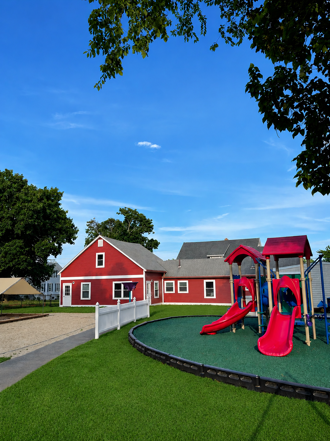 A backyard playground with a red slide and climbing structure, green grass, a white fence, and a red barn-style house under a blue sky with trees.